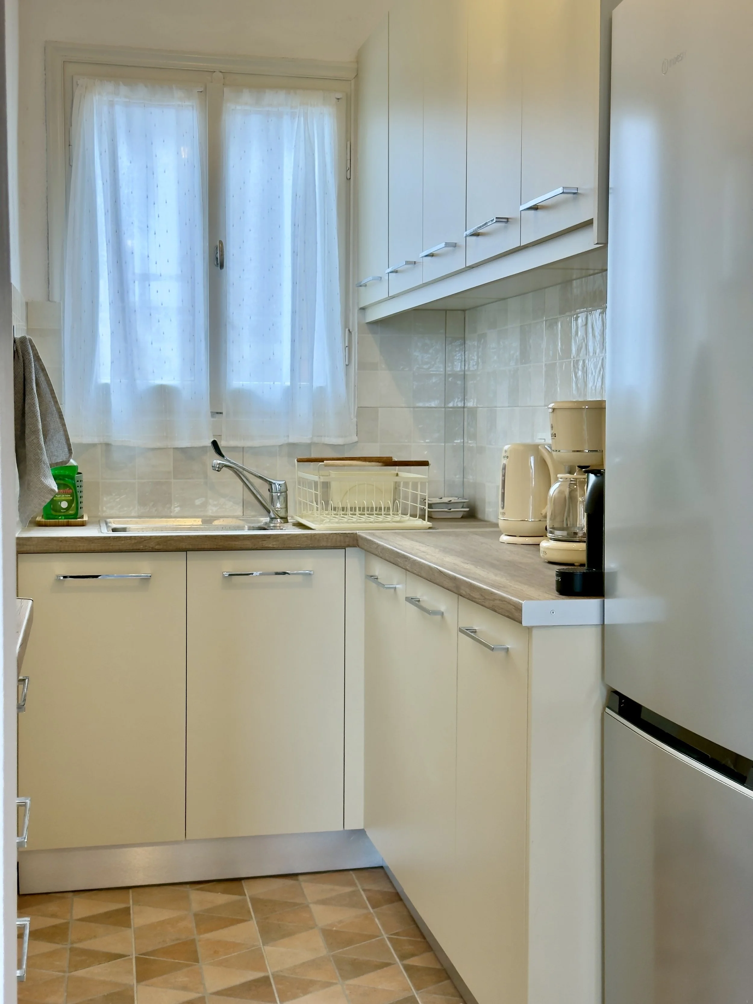 A kitchen with cream-colored cabinets, a window with sheer curtains, and a tiled floor. Items on the countertop include a dish rack, a kettle, a coffee maker, and a toaster.