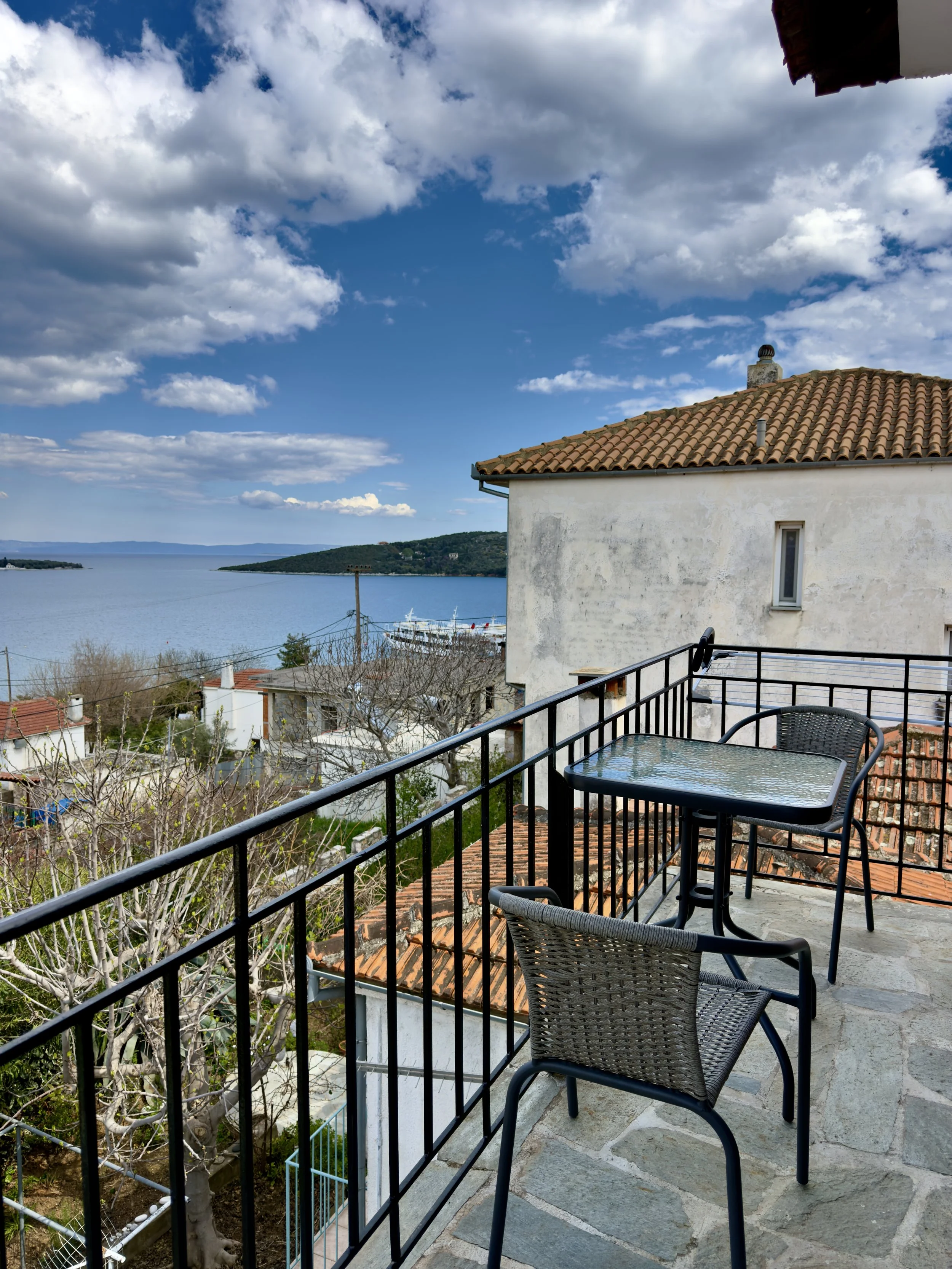 A balcony with a black metal railing overlooking a view of the water and hills in the distance. There are two chairs and a glass table on the balcony. The sky is partly cloudy with blue sky visible.