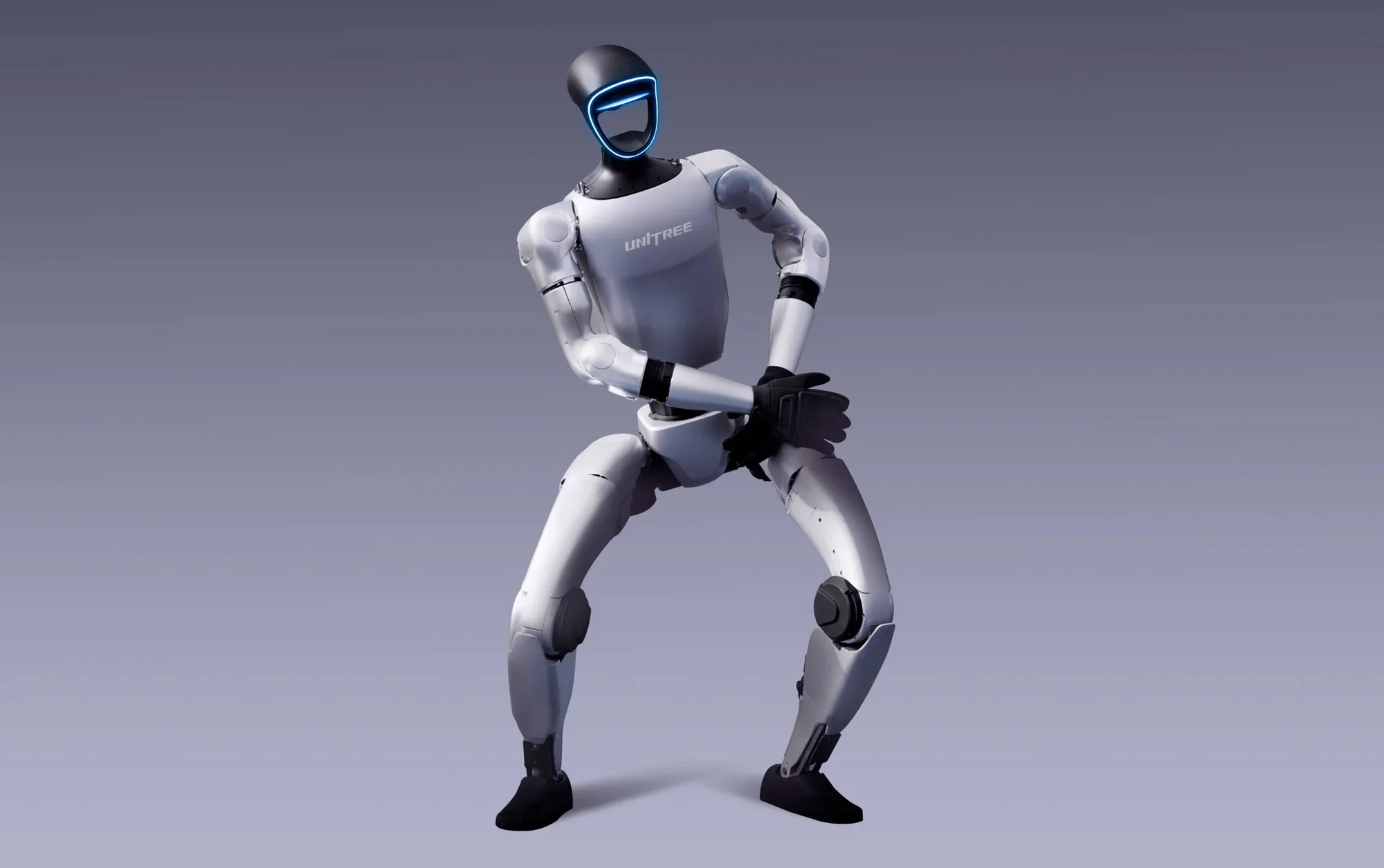 A humanoid robot with a black head and a blue LED face, wearing a gray shirt with 'UNITREE' written on it, is in a squatting position with hands clasped in front, against a plain gray background.