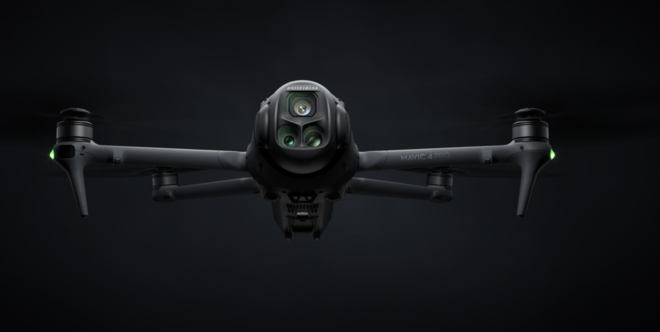 A black quadcopter drone with a camera mounted underneath, flying against a dark background.
