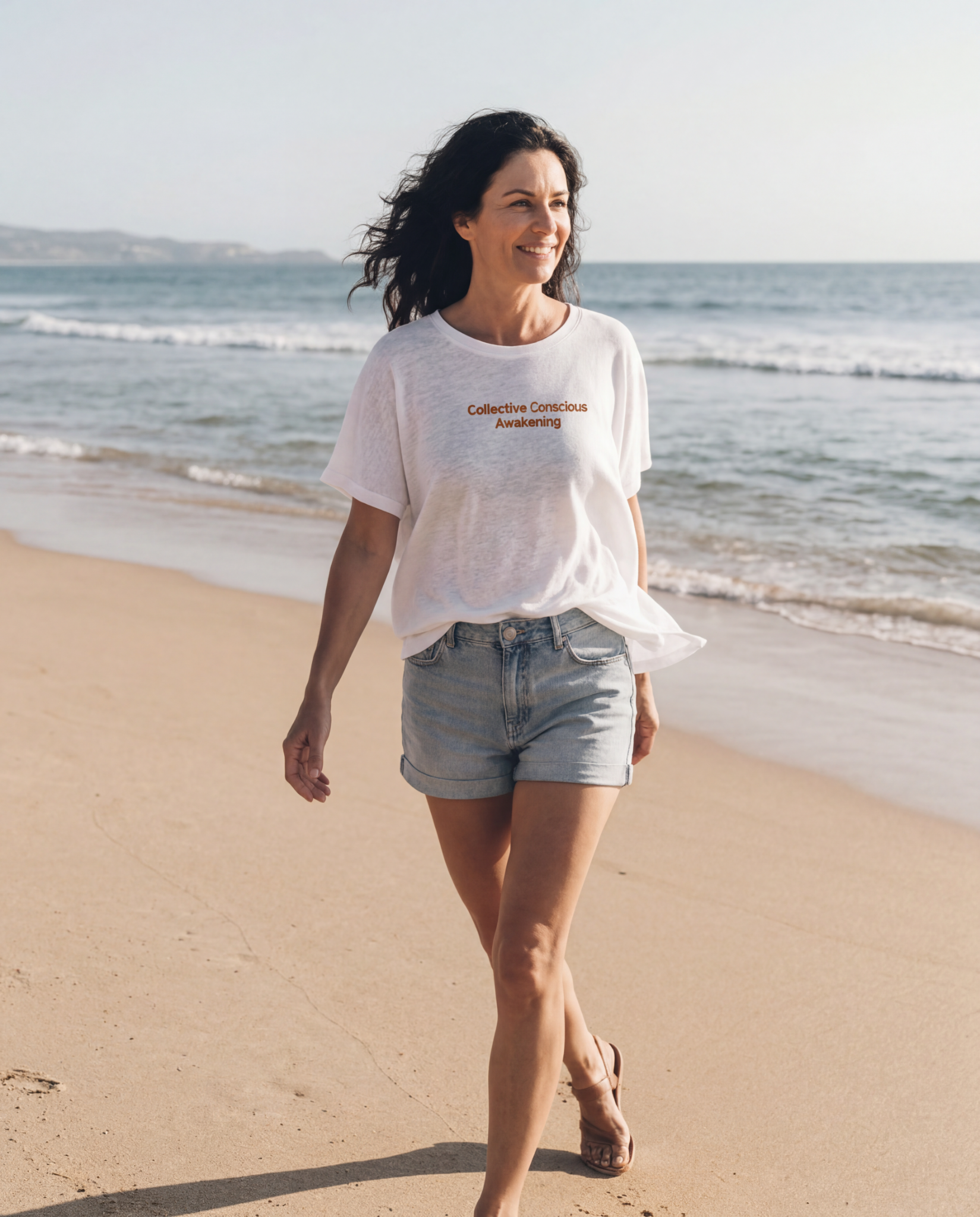 Woman walking on beach with ocean in background, wearing white t-shirt with orange text that reads 'Collective Conscious Awakening', denim shorts, and sandals, smiling and enjoying sunny weather.