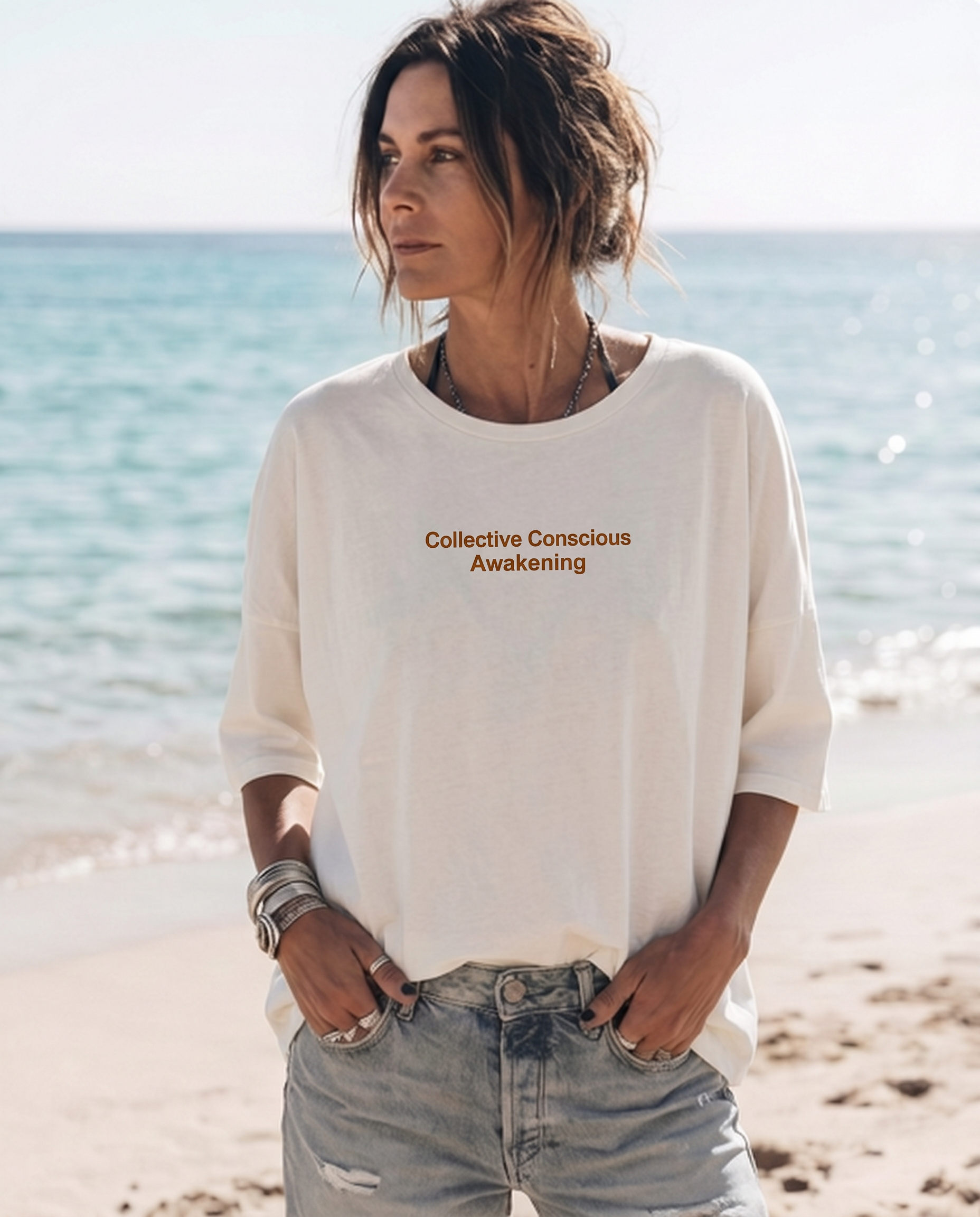 A caucasian woman standing on a beach with the ocean in the background. She has short brown hair, is wearing a white oversized sweatshirt with the words 'Collective Conscious Awakening' on it, and has her hands in her jeans pockets.