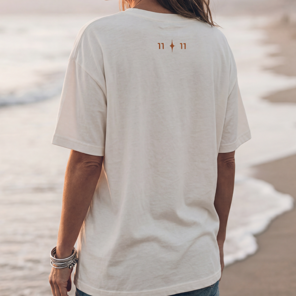 Back view of a person wearing a white T-shirt with the symbols 11:11 on the upper back, walking on the beach near the water.