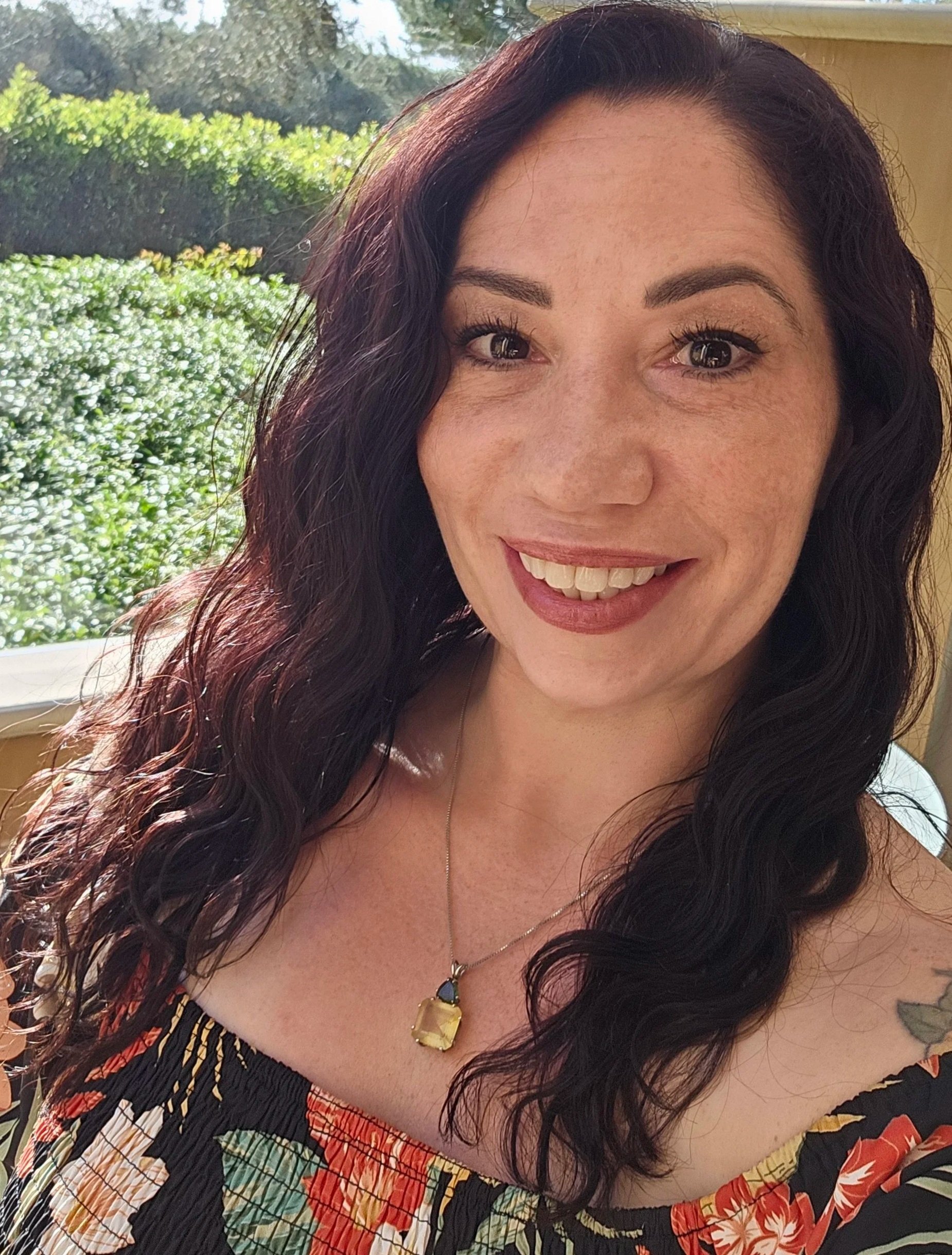 A woman with long, dark, wavy hair and a cheerful smile, wearing a colorful, patterned dress and a gold necklace with a large yellow gemstone pendant, standing outdoors near a window with greenery in the background.