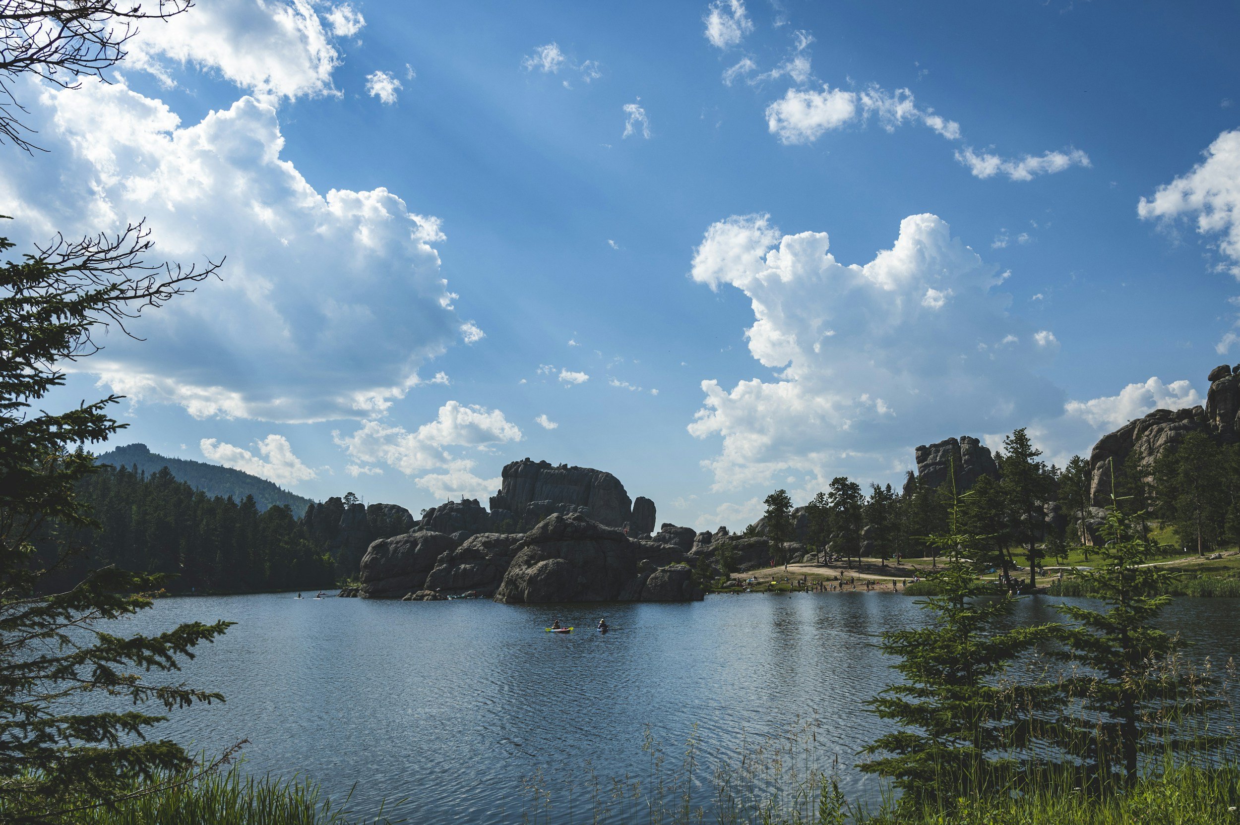 A scenic view of a lake surrounded by rocky formations and dense trees under a blue sky with scattered white clouds.