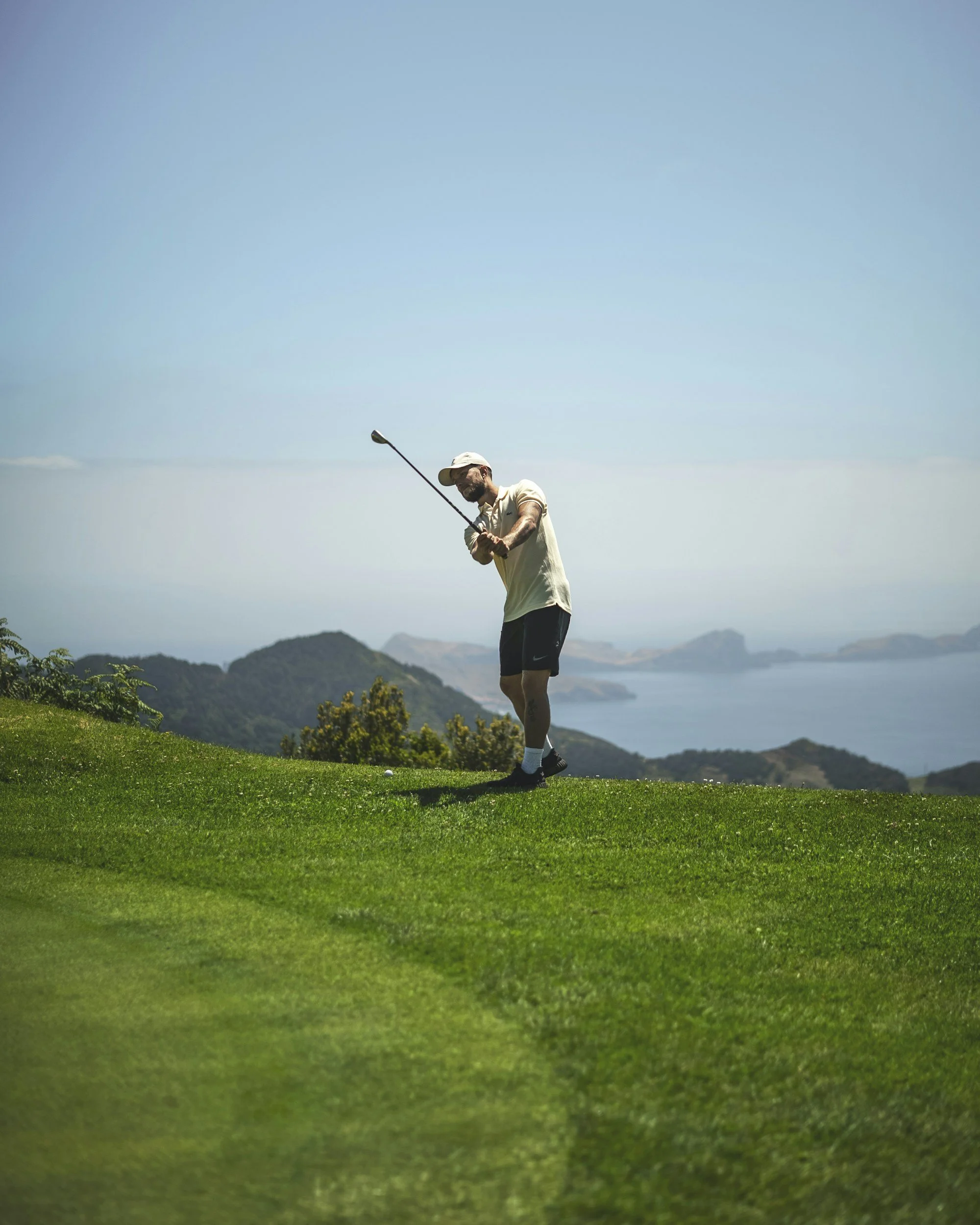 Man wearing a Cap and golf attire swinging a golf club on a lush green golf course with scenic hills and water in the background under a clear sky.