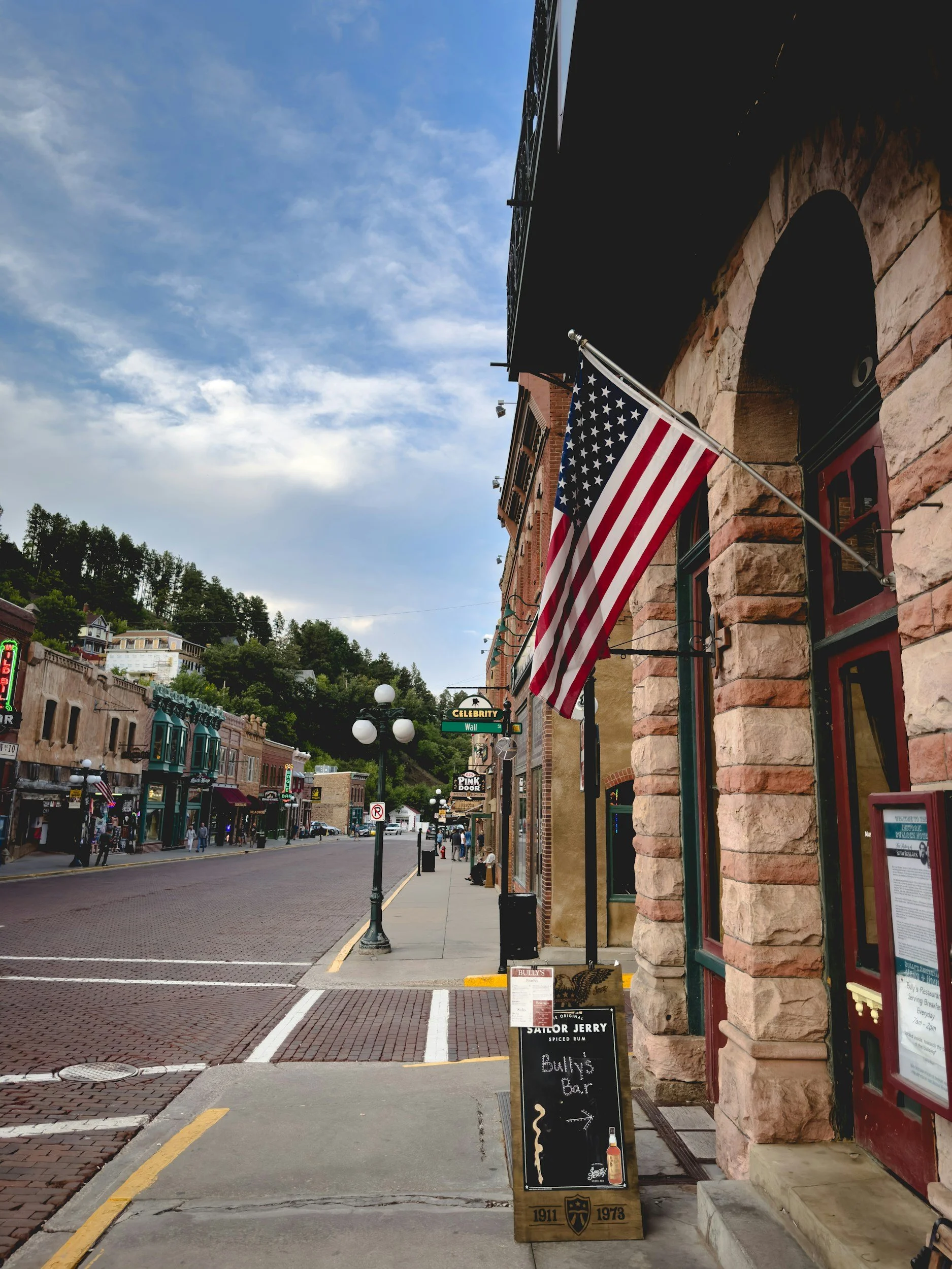 A street scene in a historic downtown area with brick and stone buildings, American flags, street lamps, and pedestrians. A chalkboard sign advertises a bar and whiskey.