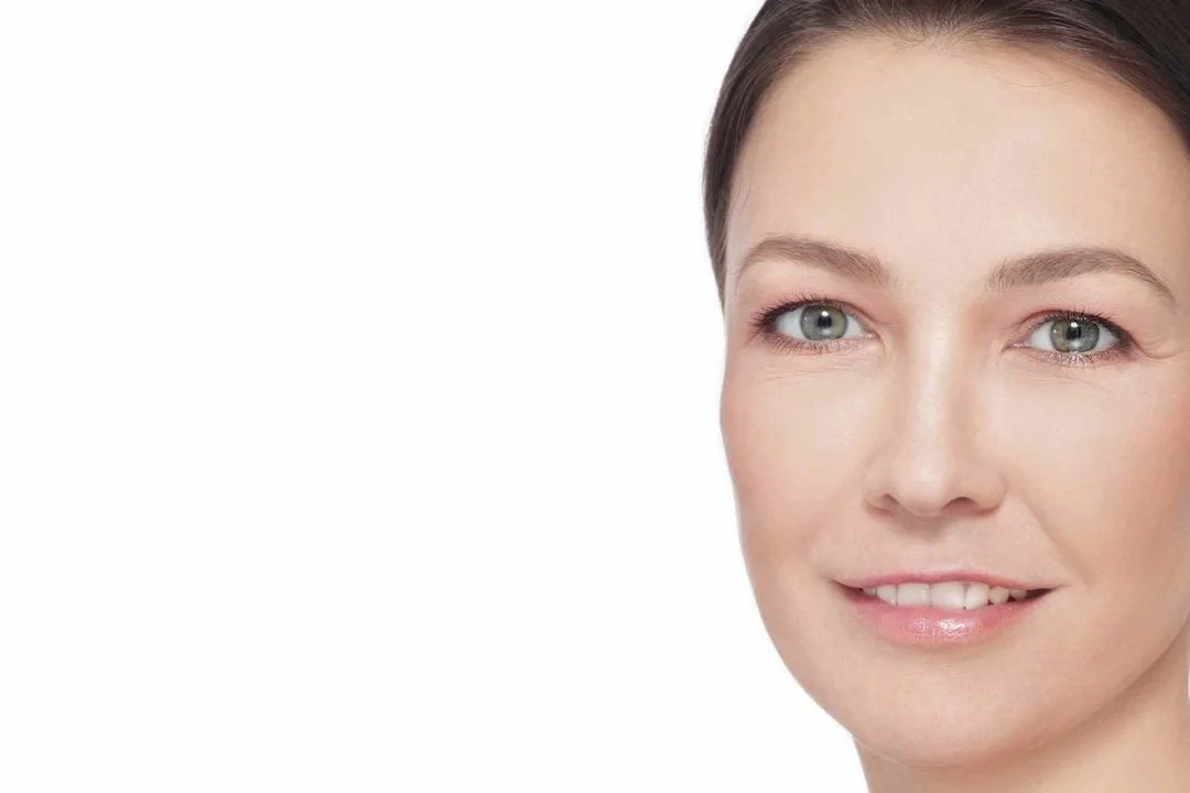 Close-up of a woman with clear skin, light makeup, and blue eyes, smiling softly against a white background.