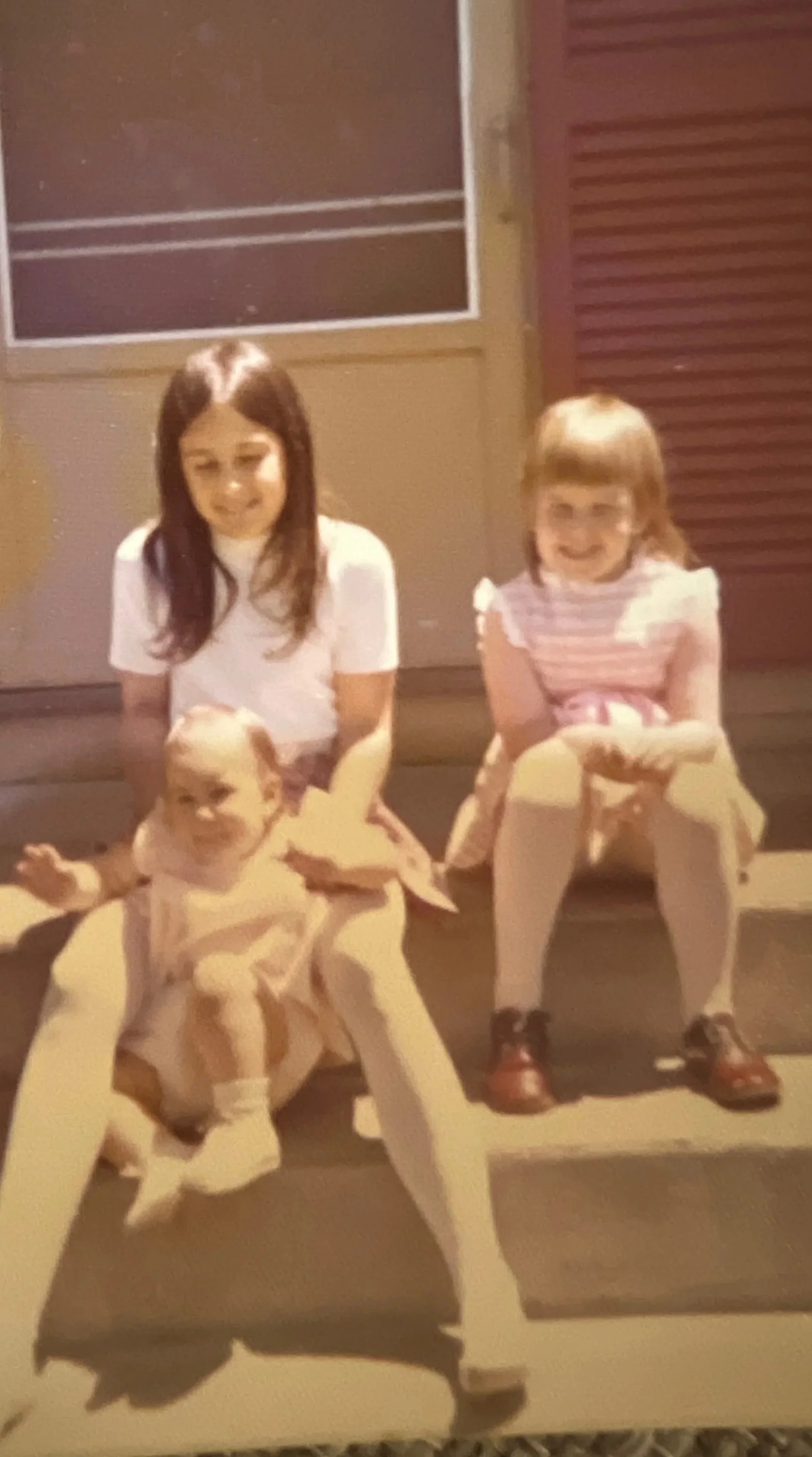 Three children sit on steps in front of a building, with one young girl in a dress with short hair, an older girl with long dark hair, and another girl with red hair and striped shirt, all smiling. The Lucky Penny Consignment East Granby, CT
