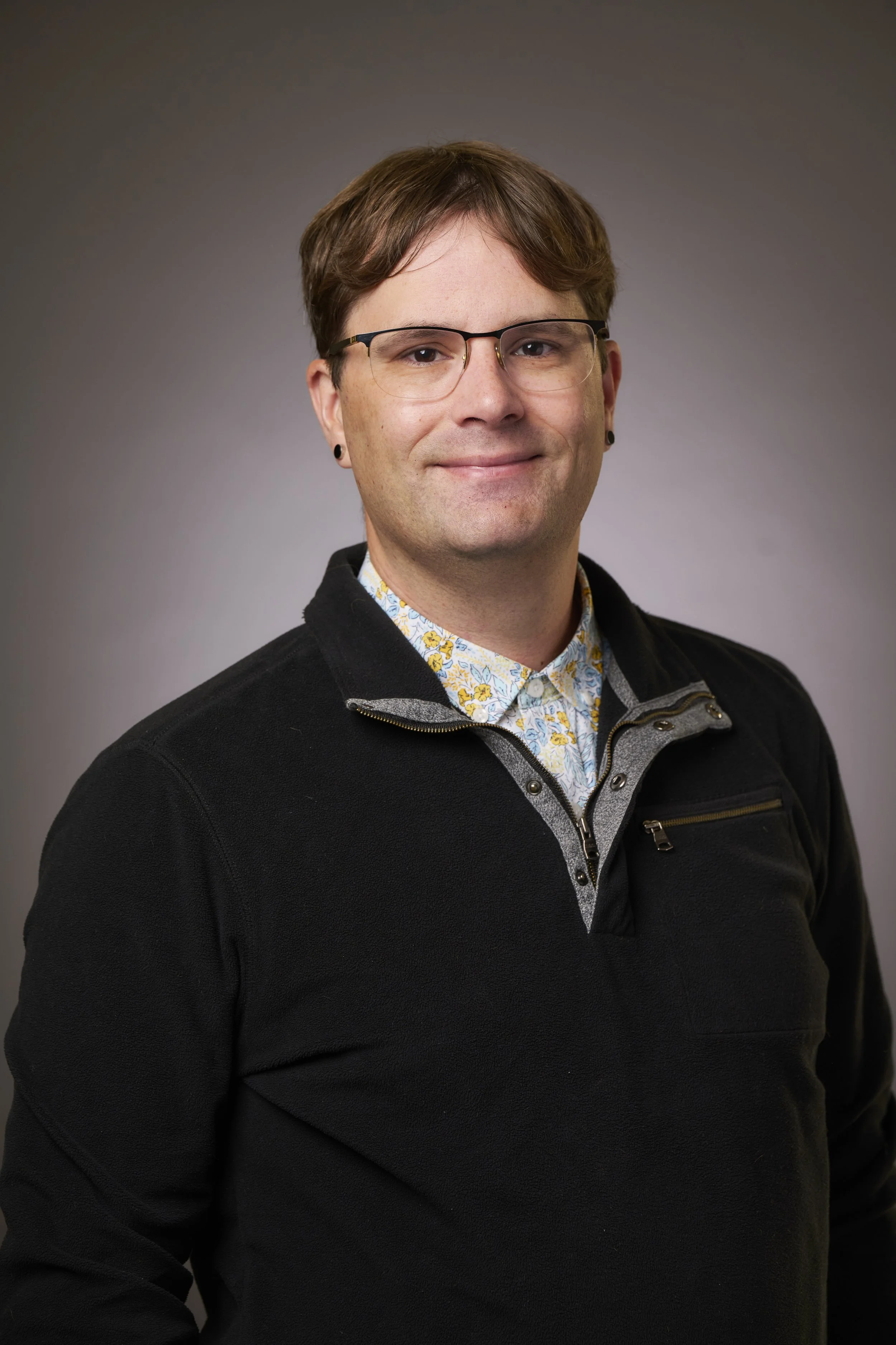 A man with light skin, brown hair, glasses, and earrings, wearing a floral shirt under a black zip-up sweater, smiling and looking at the camera against a gray gradient background.