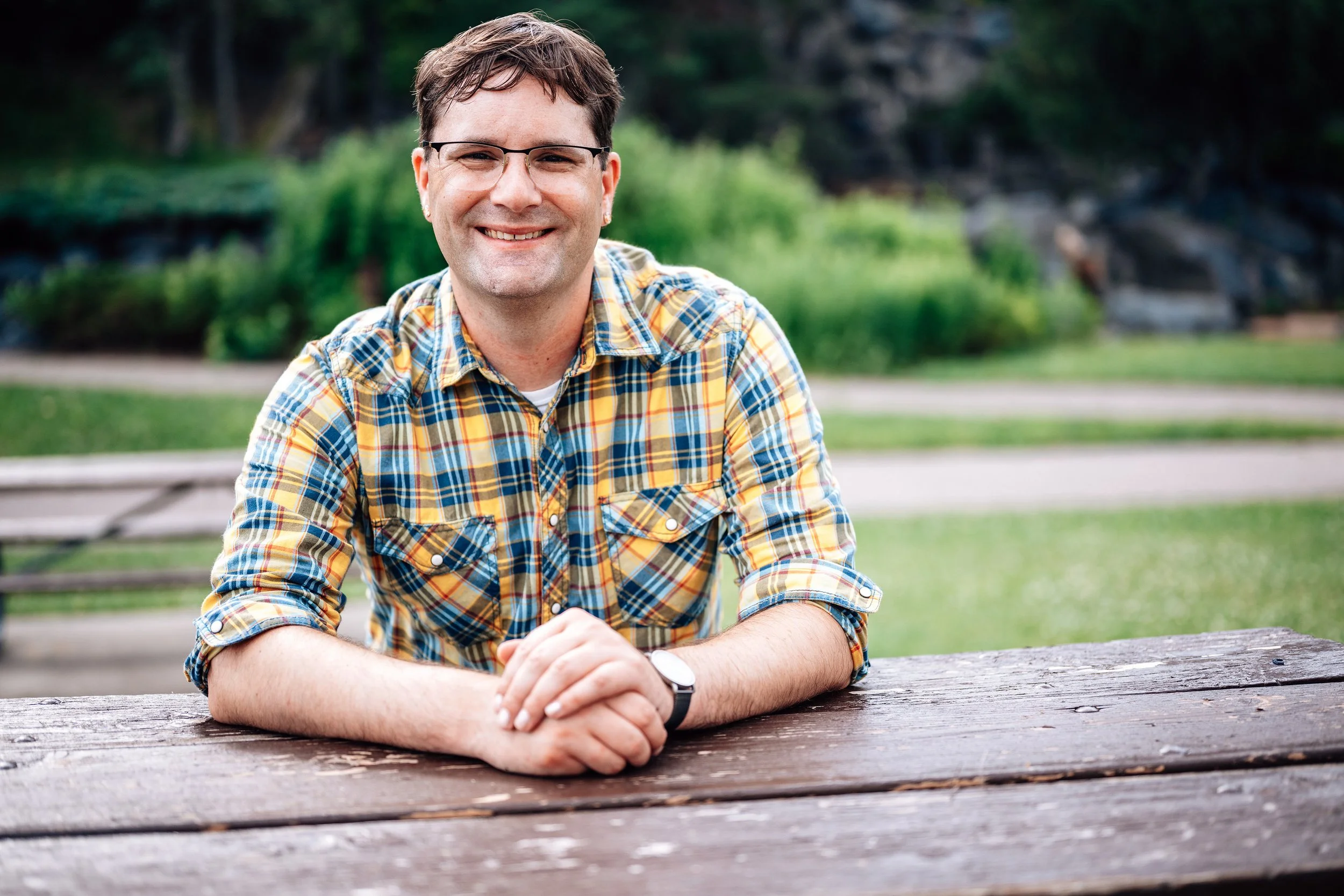 A man with short brown hair, glasses, and earrings smiling while sitting at a wooden picnic table outdoors, wearing a plaid shirt and a watch.