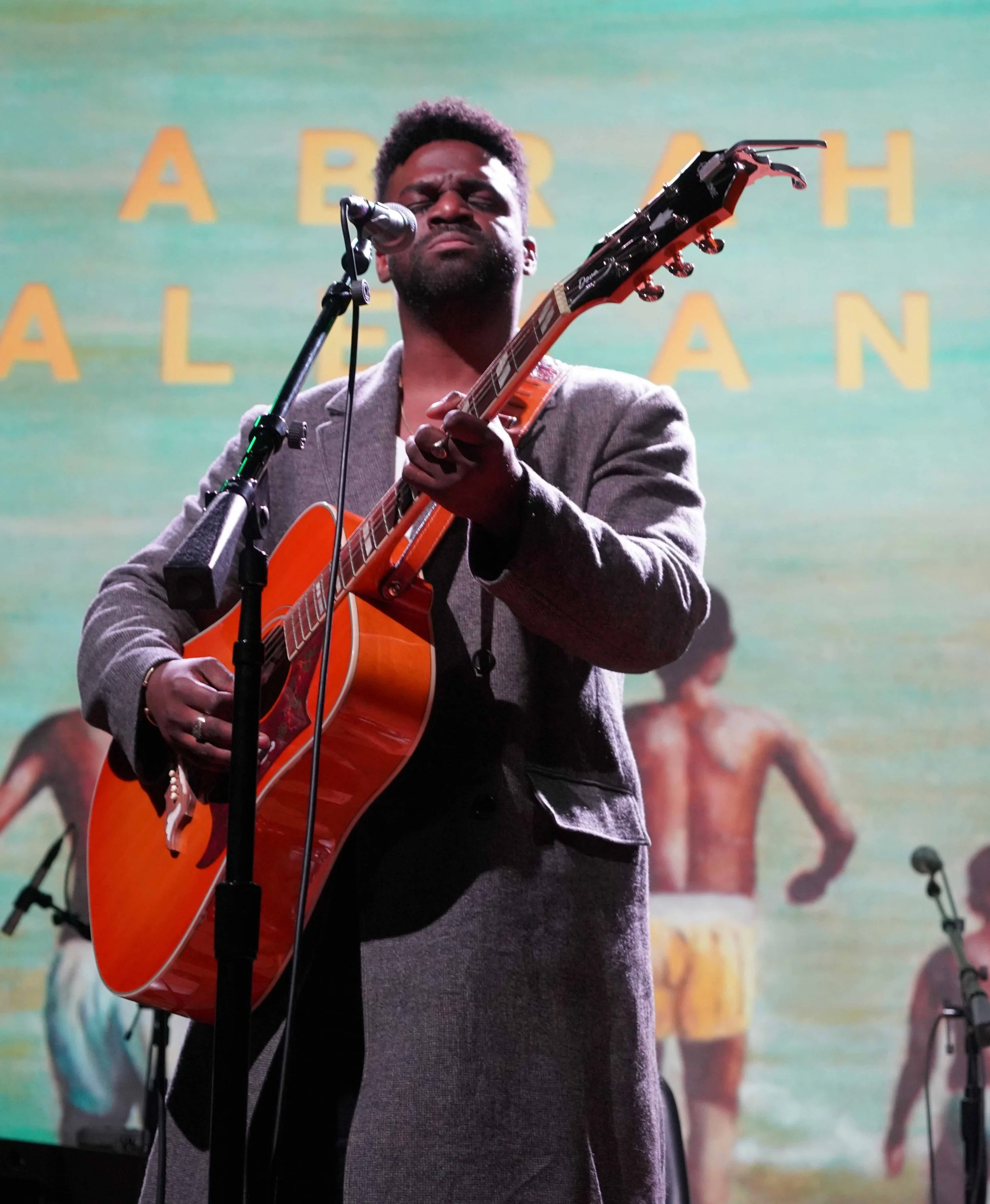 A man with dark hair and a beard passionately singing and playing an acoustic guitar on stage with a colorful beach-themed backdrop.