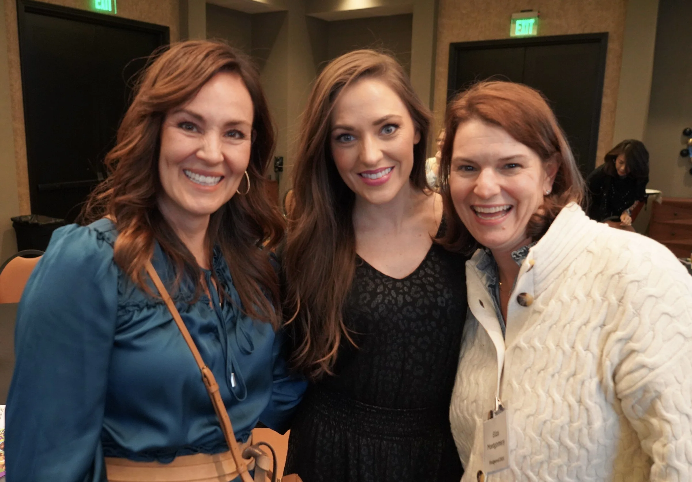Three women smiling and taking a selfie together in an indoor setting, with a woman in the background.