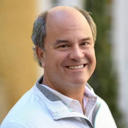 A man with gray hair in a blue sweater and beige pants poses sitting on a bench against a plain, neutral background.
