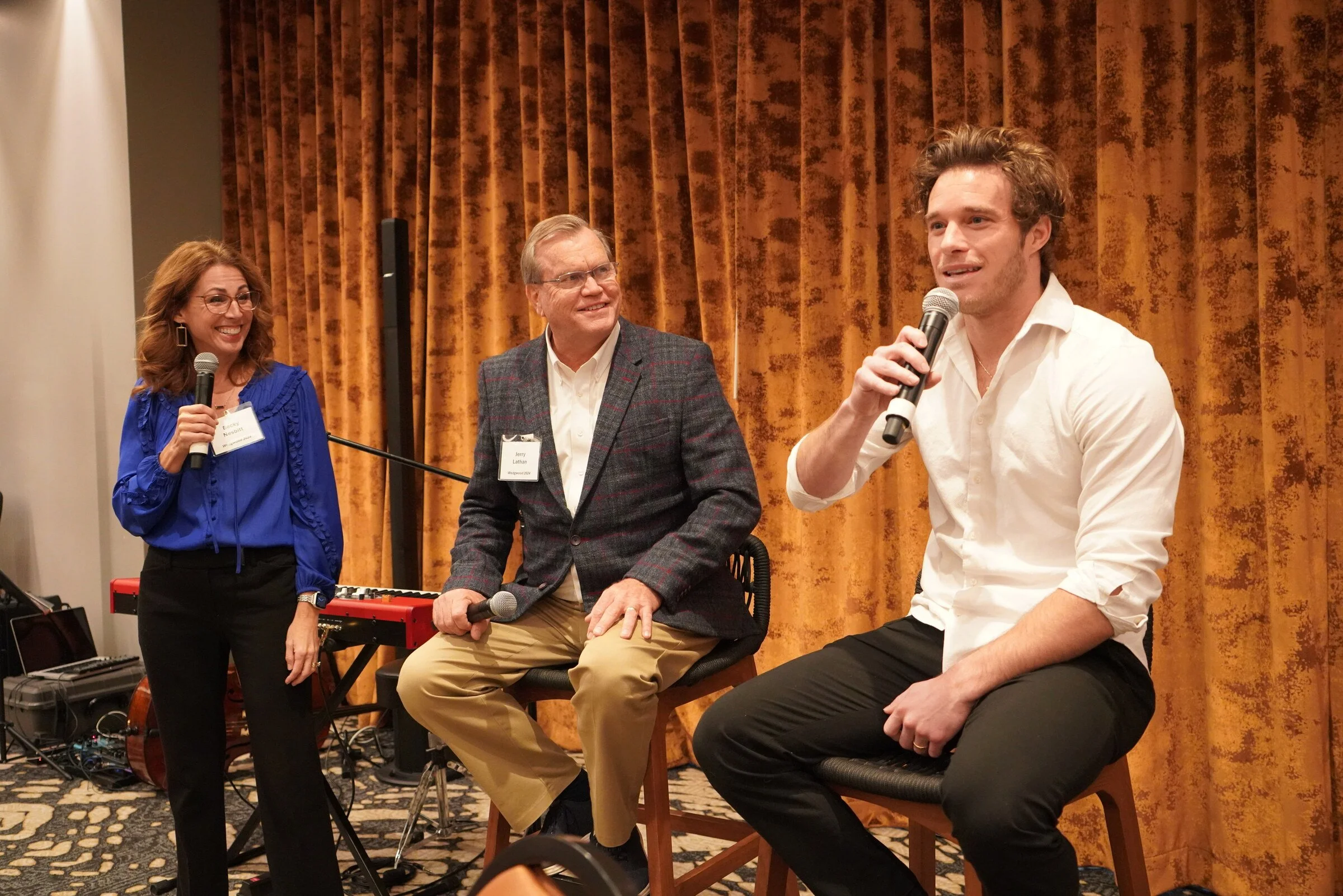 A woman and a man are standing and talking into microphones, while another man is sitting and speaking into a microphone. There are musical instruments and audio equipment in the background. The scene appears to be a discussion or event on a stage wi