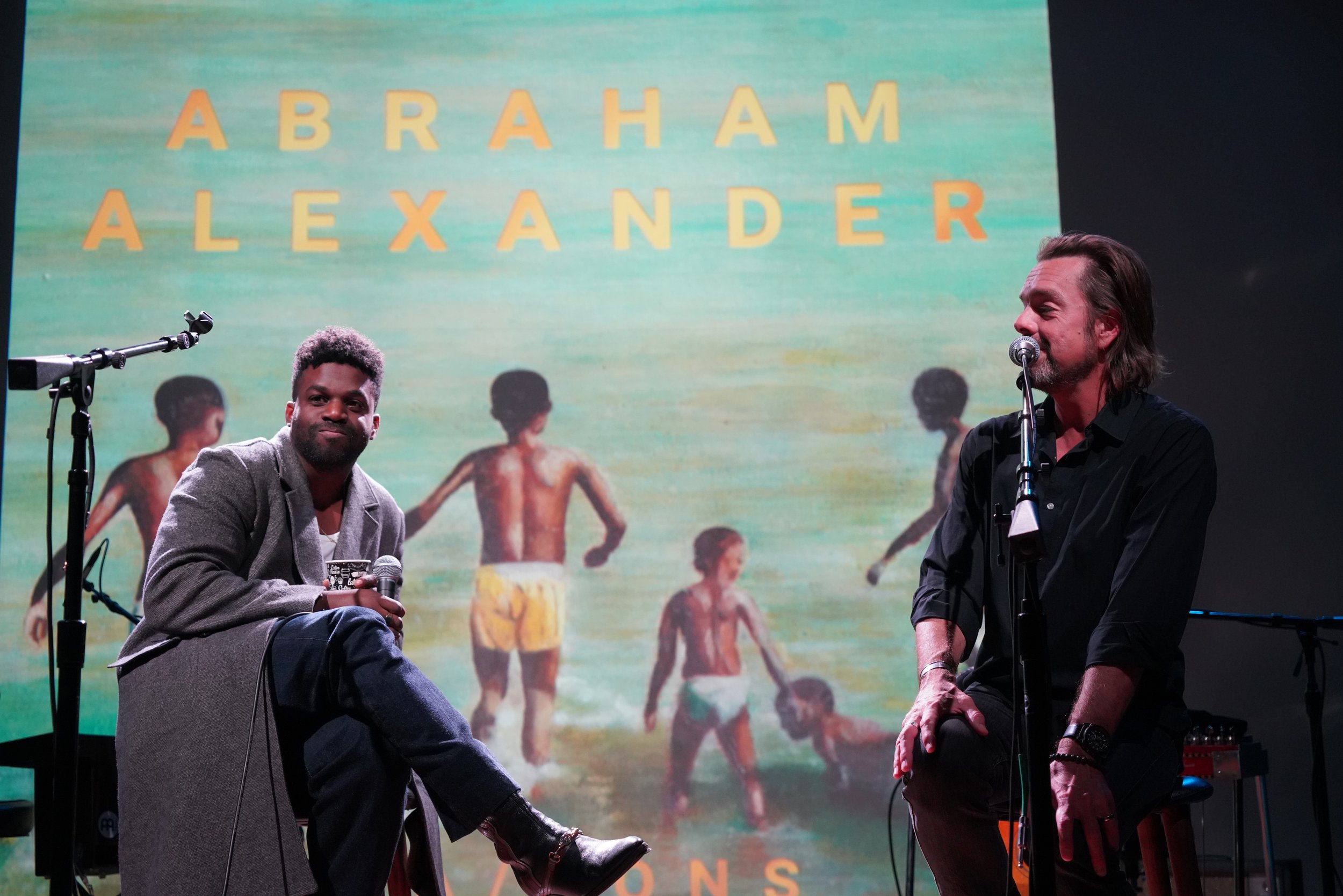 Two men sitting on stage with a large screen behind them displaying the words 'ABRAHAM ALEXANDER' and a background image of children playing outside.