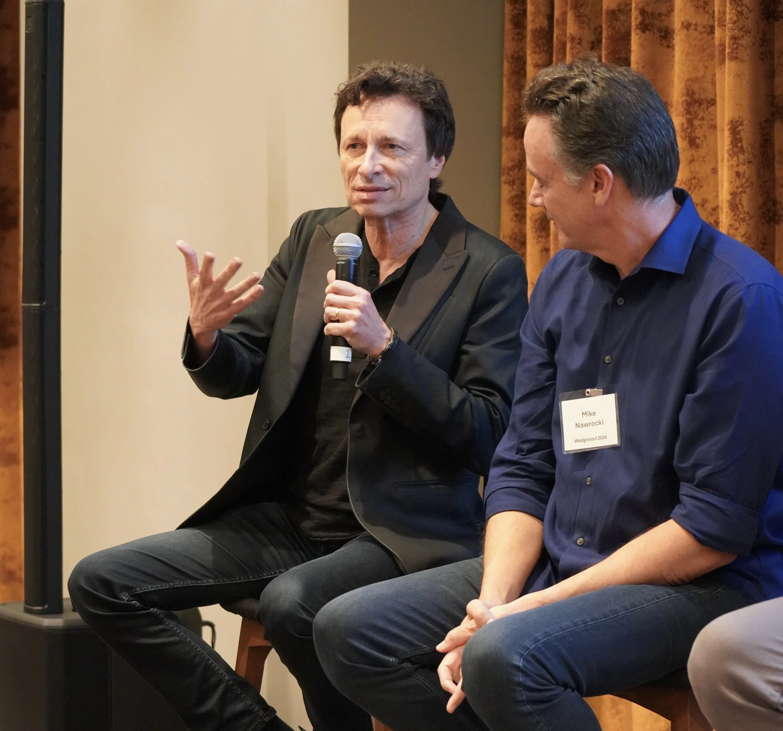 Two men sitting next to each other at a panel discussion. The man on the left is speaking into a microphone, gesturing with his hand, and looking engaged. He has dark, curly hair and is wearing a black blazer. The man on the right, with short wavy ha