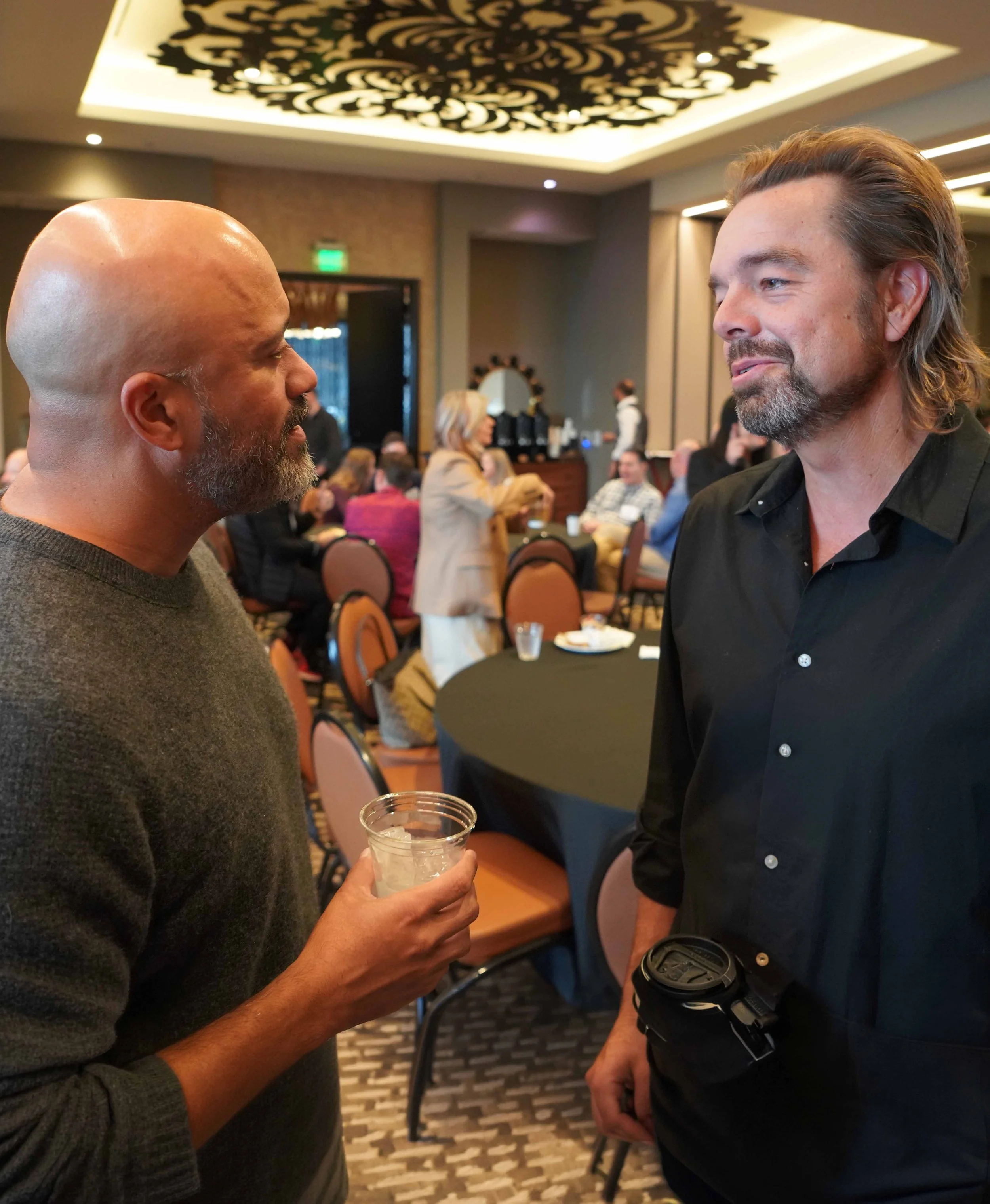 Two men engaged in conversation at a social event or conference nursing drinks, with a busy room of people and a decorative ceiling in the background.