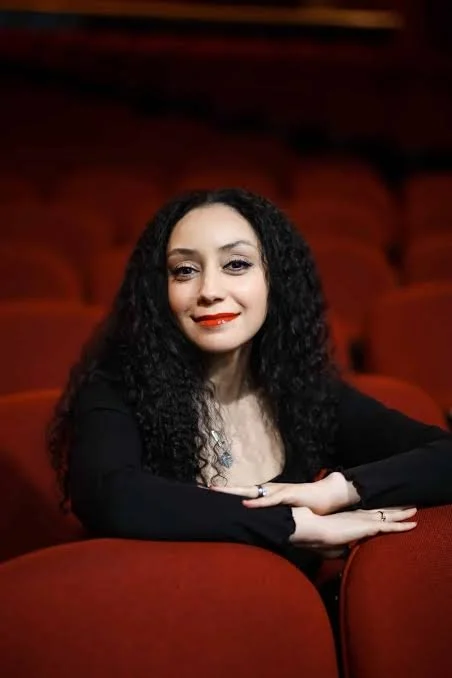 A woman with curly black hair and red lipstick sitting in a theater with red seats.