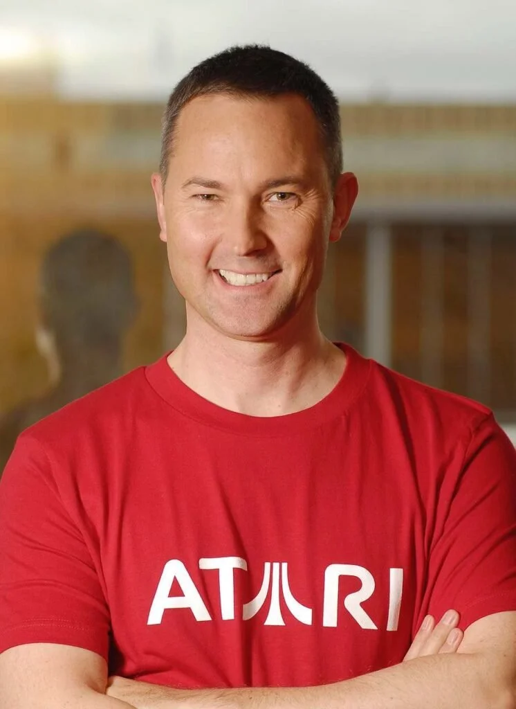 Smiling man with short dark hair wearing a red t-shirt with white letters, arms crossed, standing outdoors.