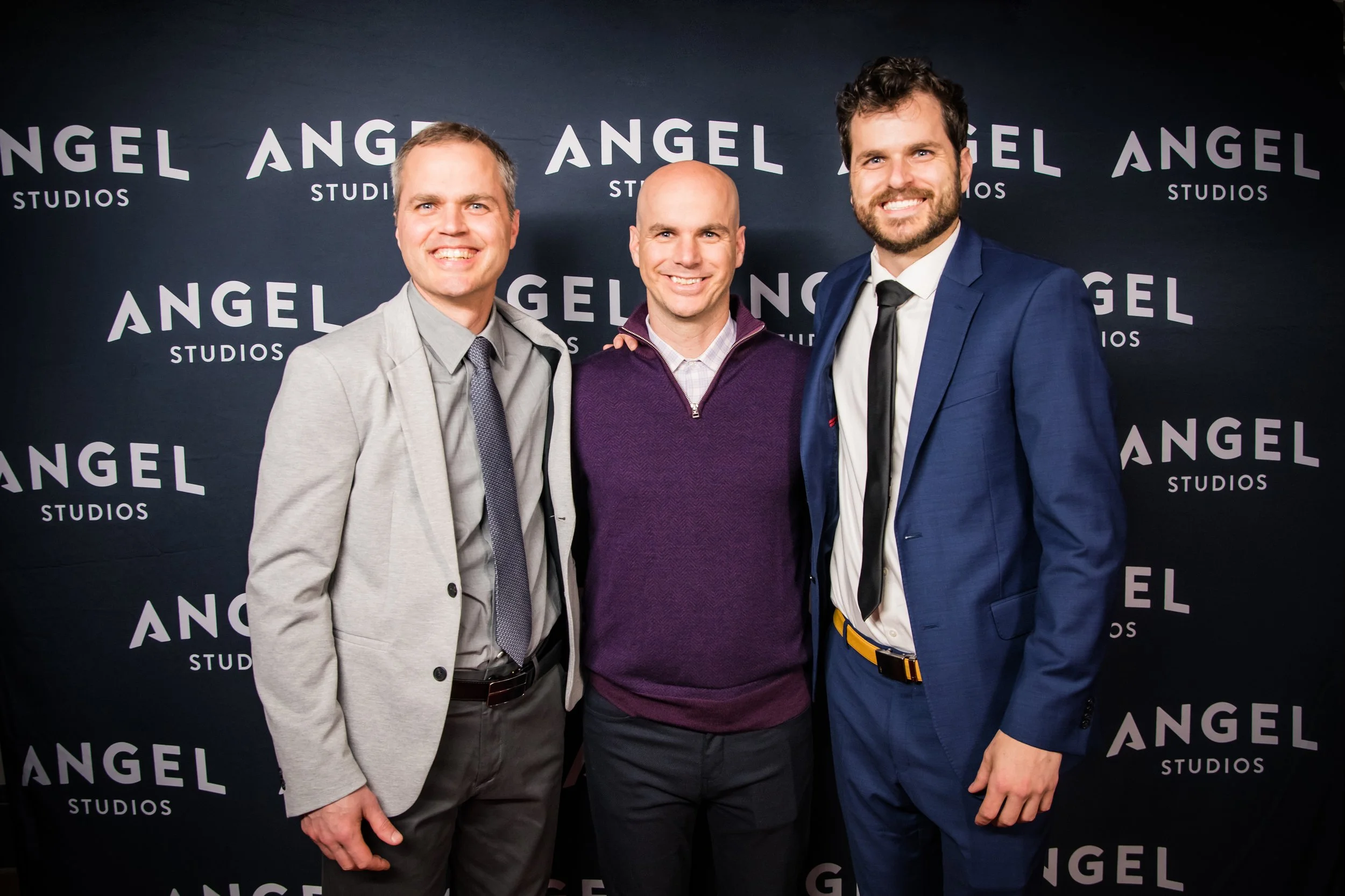 Three men smiling and standing close together in front of a black background with white text reading 'ANGEL STUDIOS' repeatedly. The man on the left is wearing a light gray blazer, the man in the middle is wearing a purple sweater, and the man on the right is wearing a blue suit with a black tie.