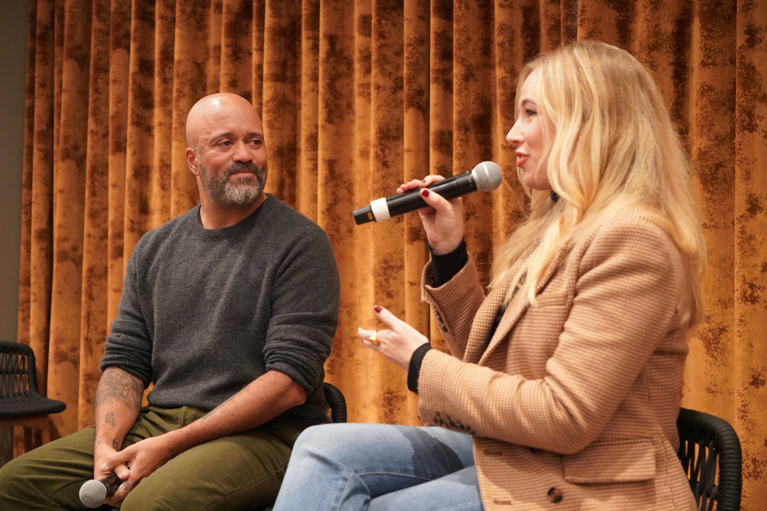 A woman with long blonde hair speaking into a microphone during an event, with a man with a bald head and gray beard listening attentively, both seated against a background of brown curtains.