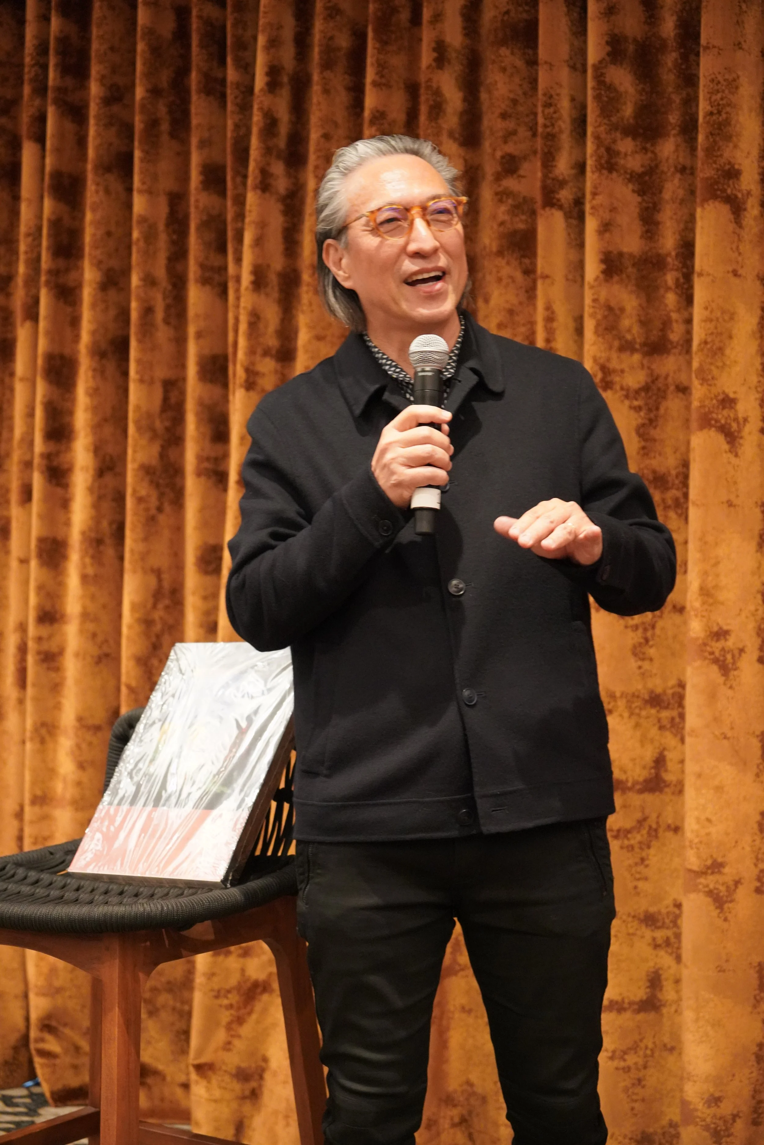 A man with gray hair and glasses holding a microphone, speaking in front of an orange velvet curtain.