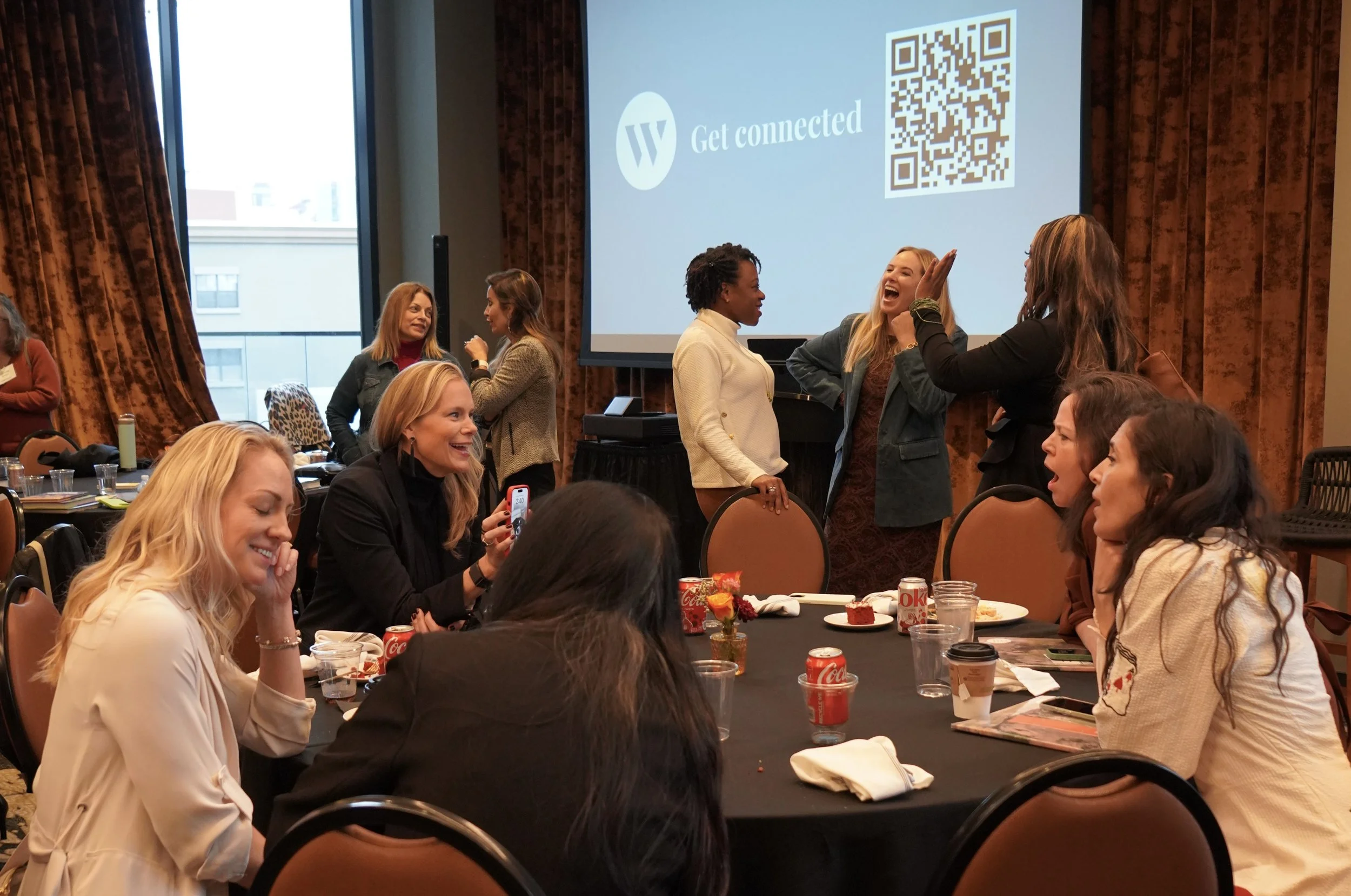 Group of women at the Wedgwood Circle Annual Event, engaging in conversation, sitting and standing around a round table with drinks and snacks in a banquet hall, with a large screen displaying a QR code and the message 'Get connected'.