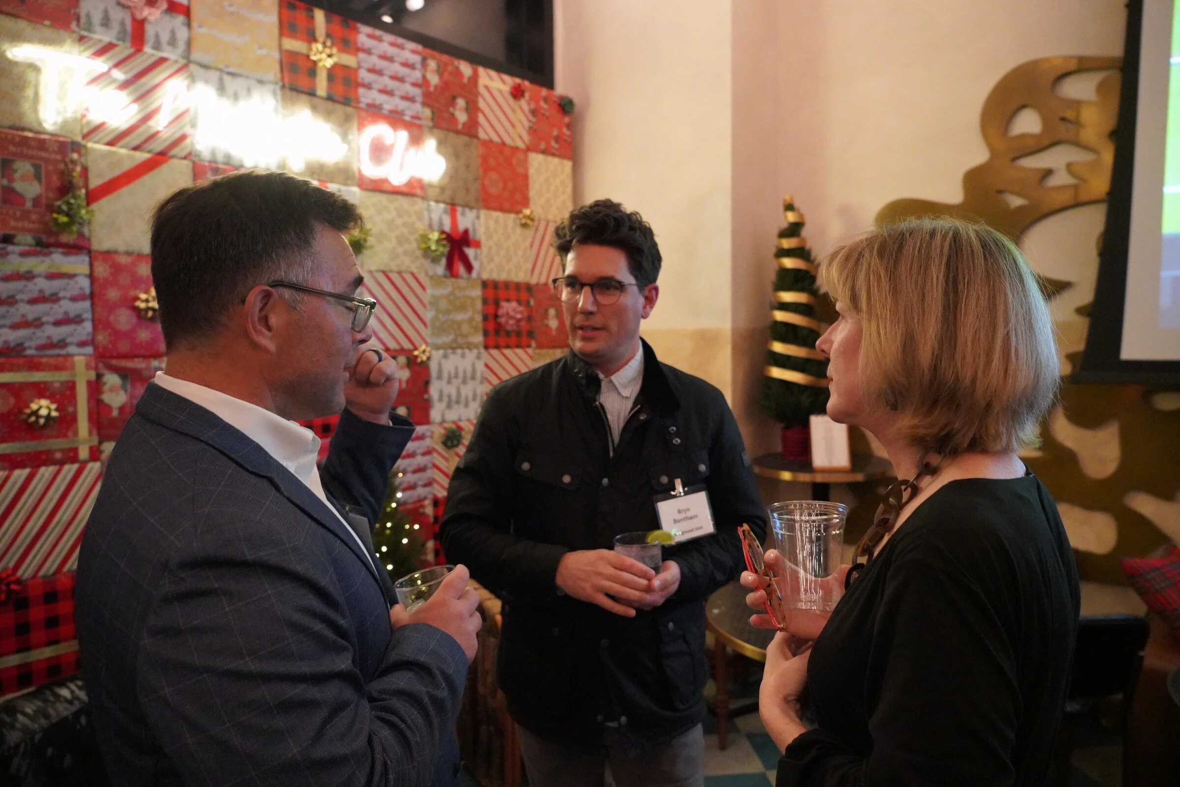 Three people talking at a holiday gathering with Christmas decorations and lights in the background.