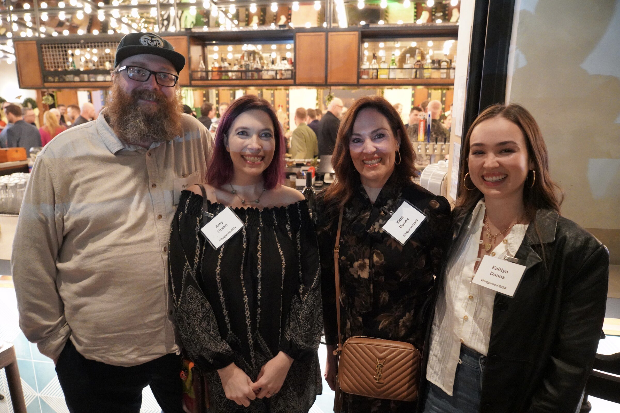 Four people standing together at an indoor event, smiling for the camera. The background shows a crowded bar or restaurant with many patrons and hanging lights.