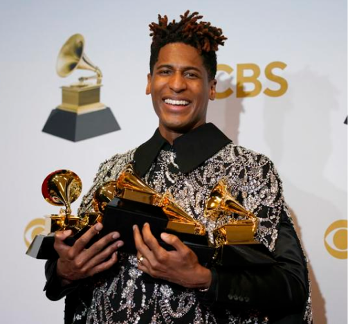 Jon Batiste smiling while holding multiple Grammy Awards, standing in front of a backdrop with the CBS logo and Grammy trophy images.