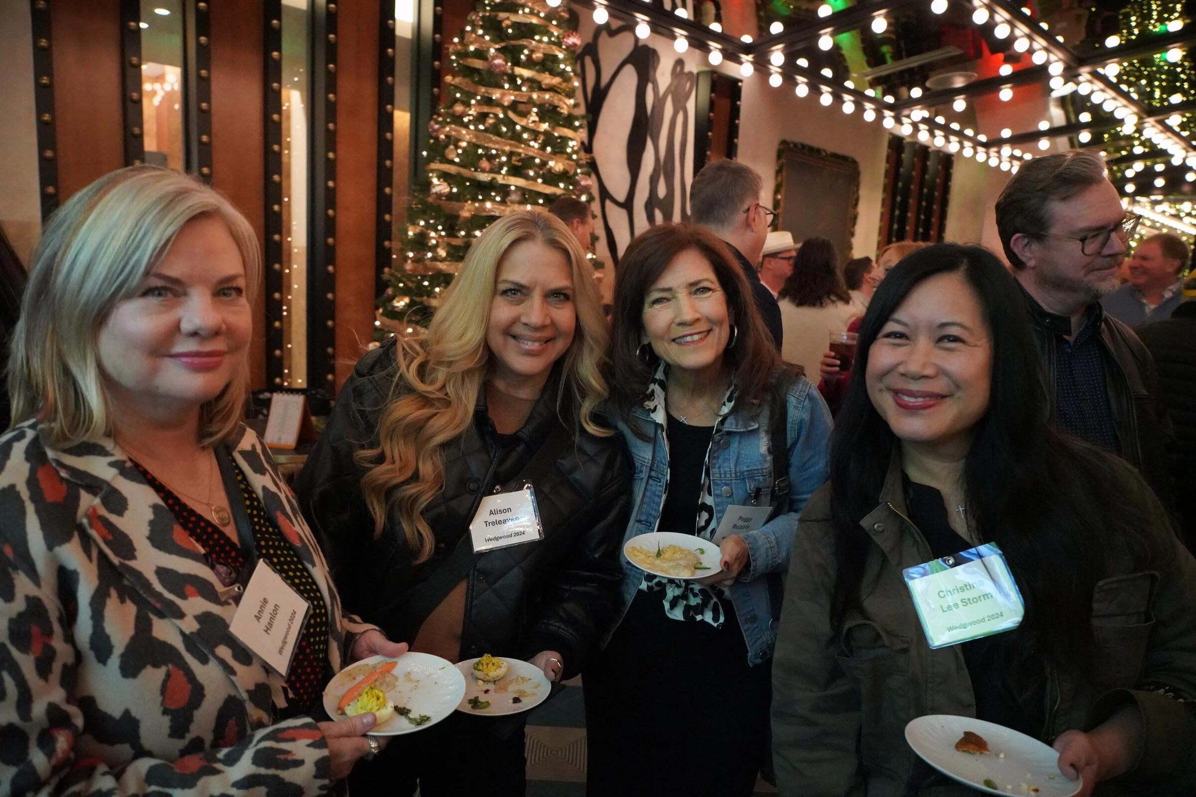 Group of people smiling and enjoying food at a festive holiday gathering with string lights and a decorated Christmas tree in the background.