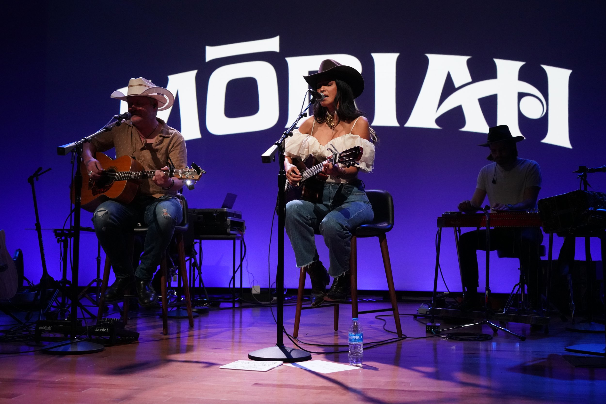 Three musicians performing on stage with a large backdrop displaying the word 'MOTEX'. The middle musician is a woman wearing a cowboy hat, playing a guitar, and singing into a microphone. The musician on the left is a man wearing a cowboy hat, playi