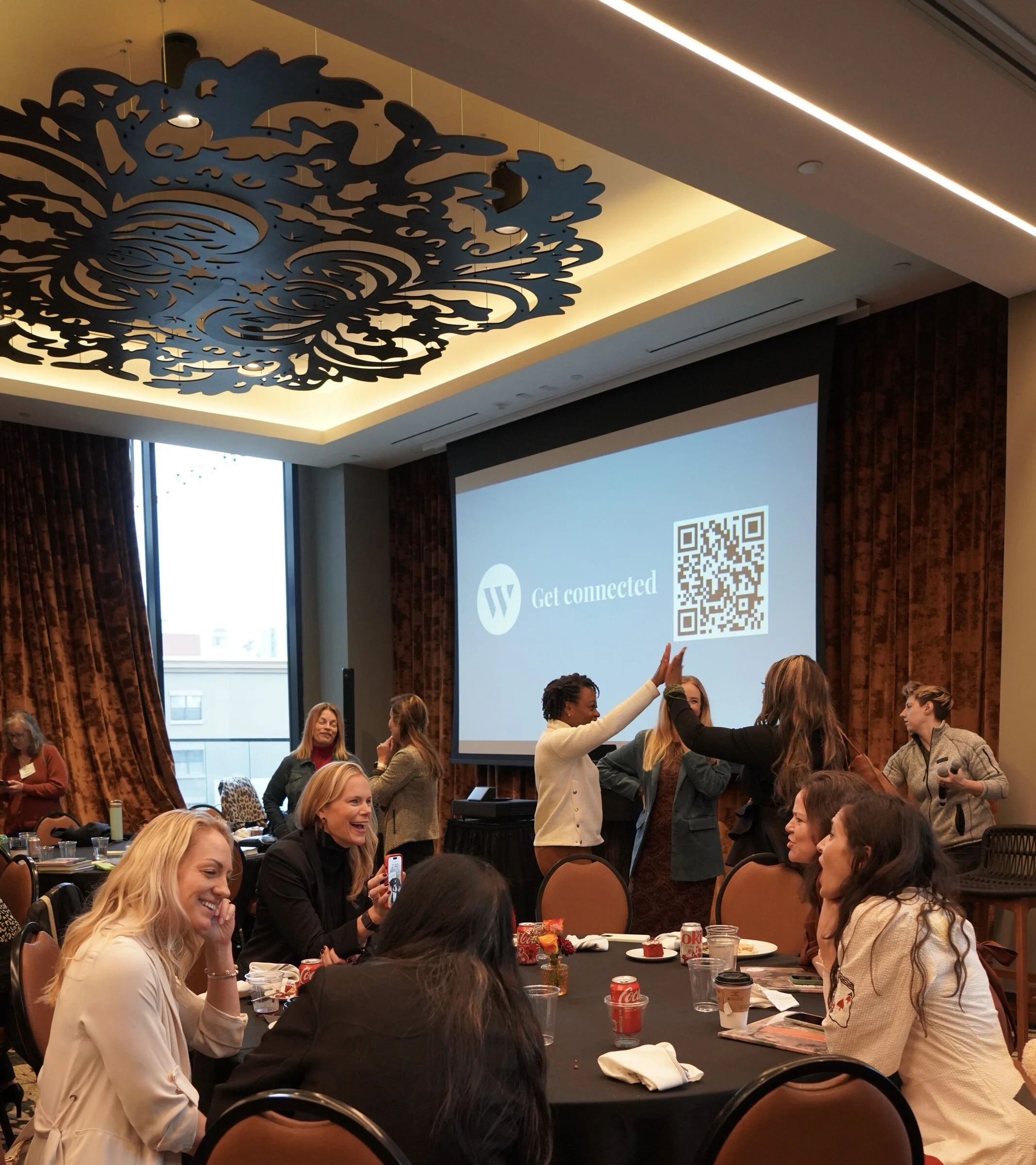 A group of women and men gathered around tables in a conference room, with one woman and one man standing and fist-bumping near the large projection screen displaying a QR code. The room has large windows, dark curtains, and a decorative ceiling fixt