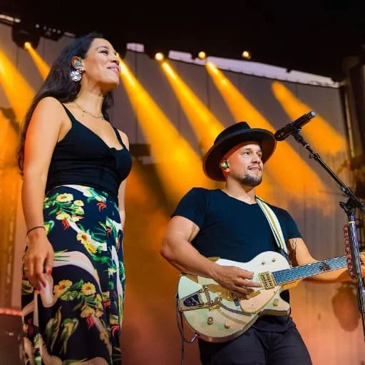A woman singing on stage with a man playing guitar, stage lights in the background.