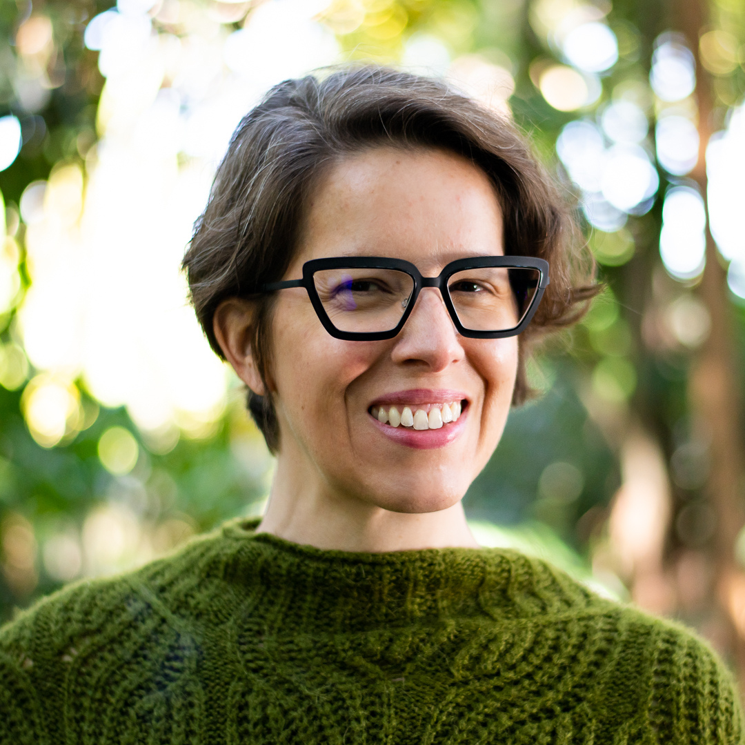 A woman smiling outdoors wearing black glasses and a green knitted sweater, with a background of blurred green trees and sunlight.