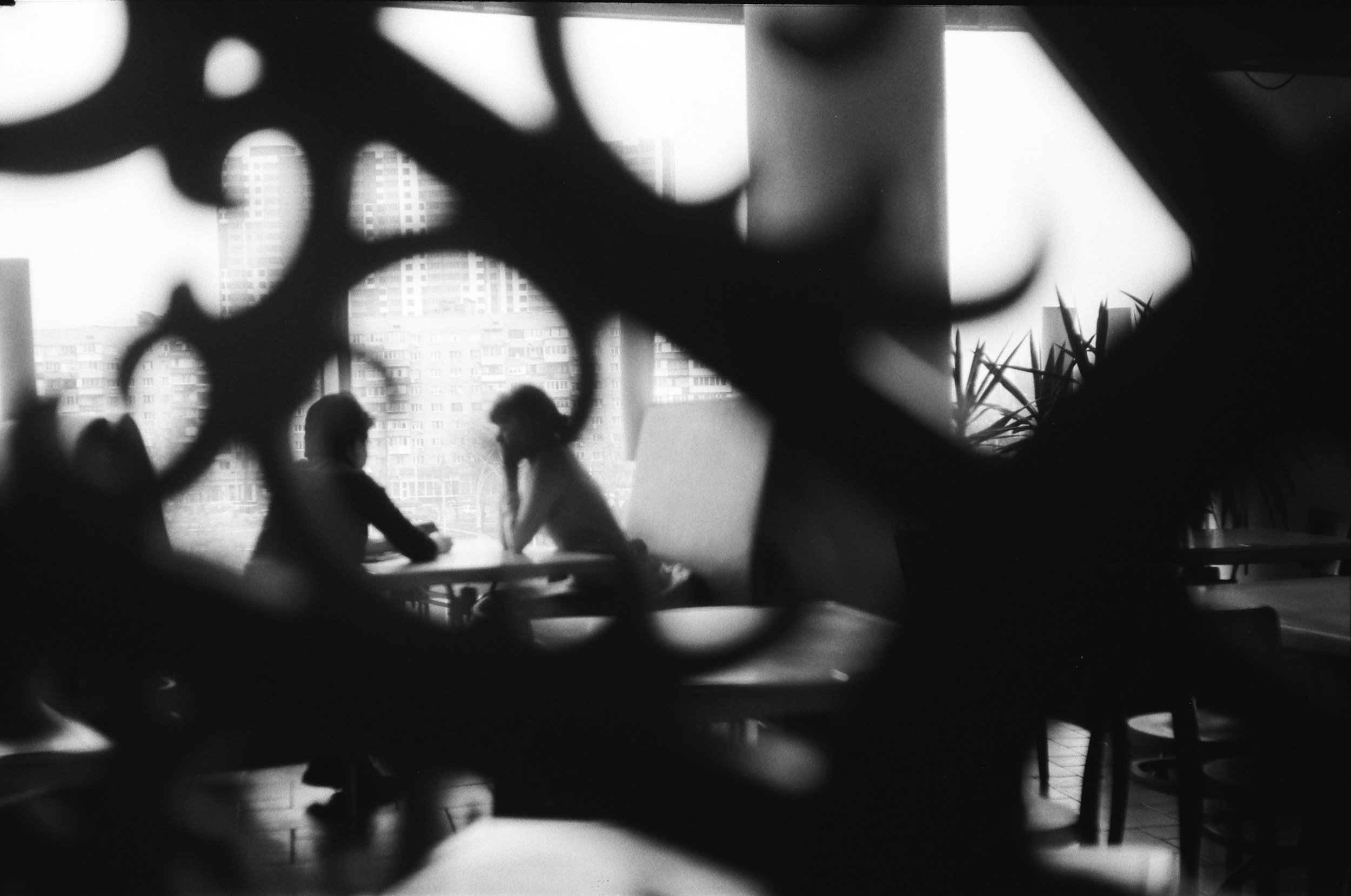 Black and white photograph of two people sitting at a table, seen through a decorative metal or wooden partition, with a background of high-rise buildings outside.