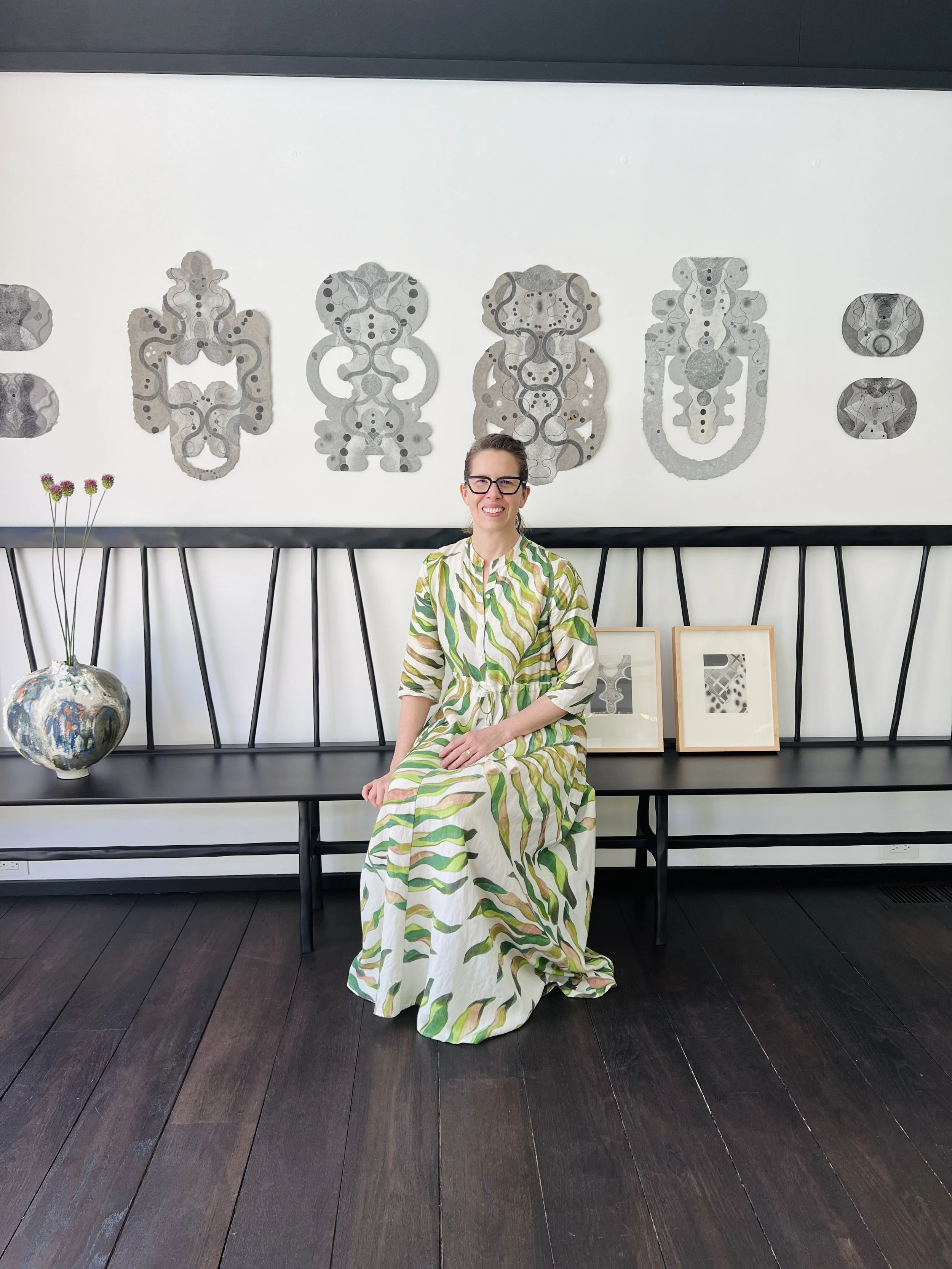 Woman wearing a green and cream botanical patterned dress sitting on a black wooden bench on a dark wooden floor.