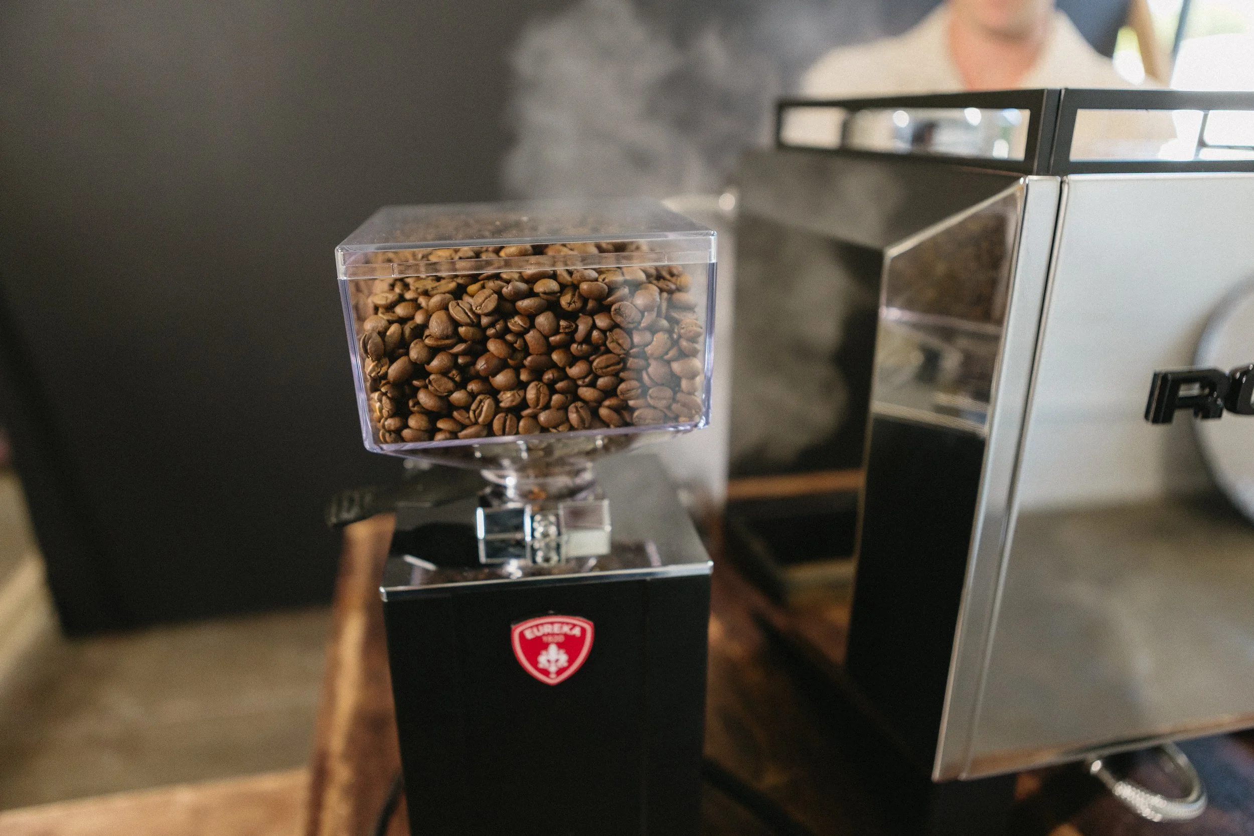 Close-up of a coffee bean grinder with a transparent container filled with roasted coffee beans. A person in the background is partially visible, operating the machine.