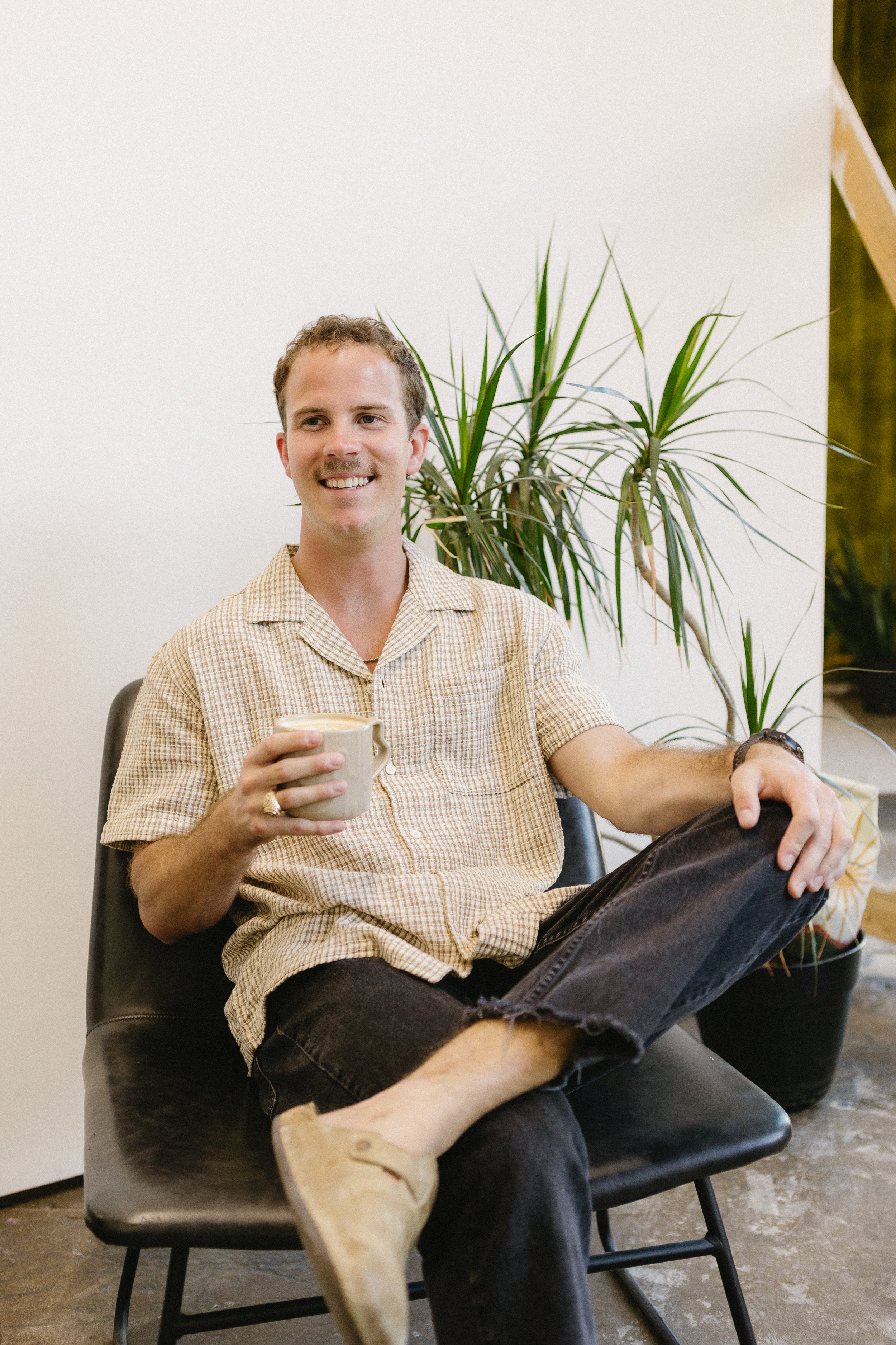 A young man with light skin and curly hair is sitting on a black chair, smiling while holding a ceramic mug. He is wearing a beige checkered shirt, black jeans, and light-colored shoes. Behind him, there is a large green plant with long leaves and a white wall. Part of a wooden structure is visible on the right side.