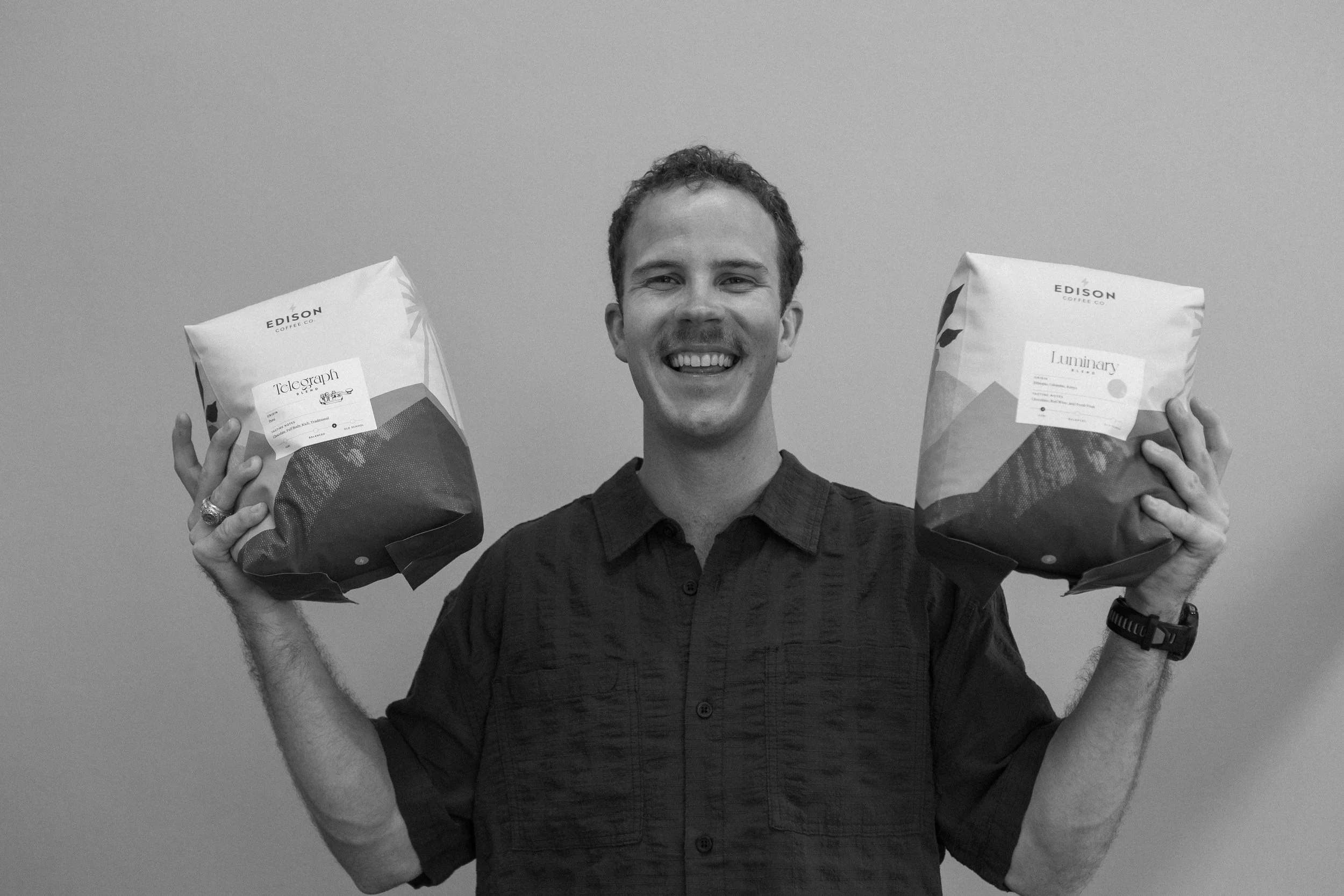 A smiling man holding two bags of coffee, one in each hand, in front of a plain background.