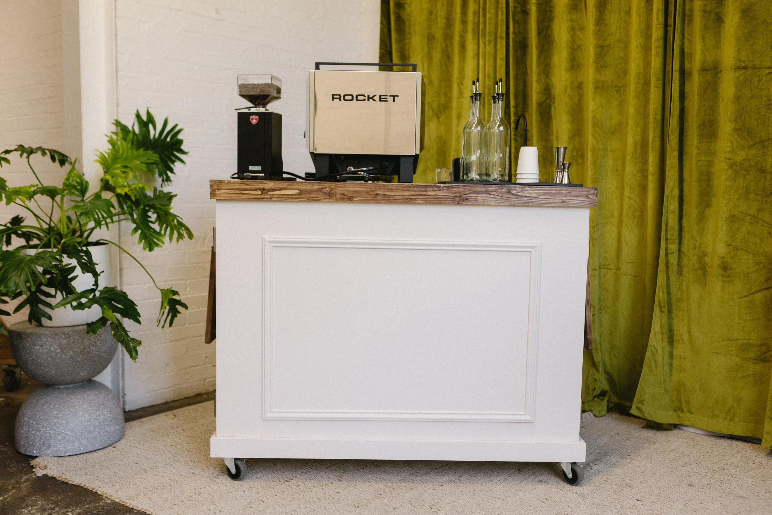 A small white counter on wheels with a wood top, set against a yellow curtain and white brick wall. To the left, in front of the counter, is a potted plant with large green leaves. On the counter are a coffee machine, glass bottles, utensils, and cups.