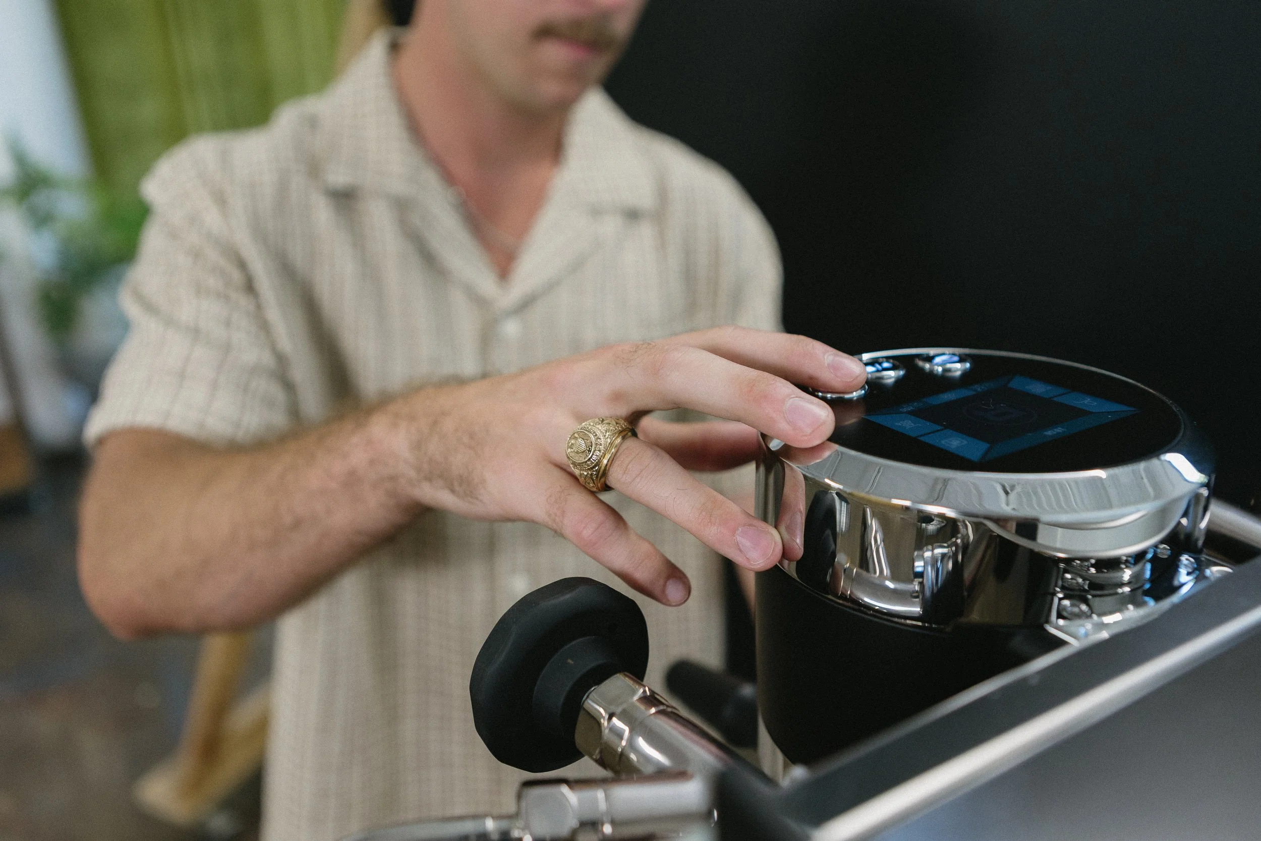 A person with a beige checkered shirt, gold ring, and mustache is adjusting or inspecting a motorcycle. The focus is on their hand and the motorcycle's dashboard, with the person's face partially visible.