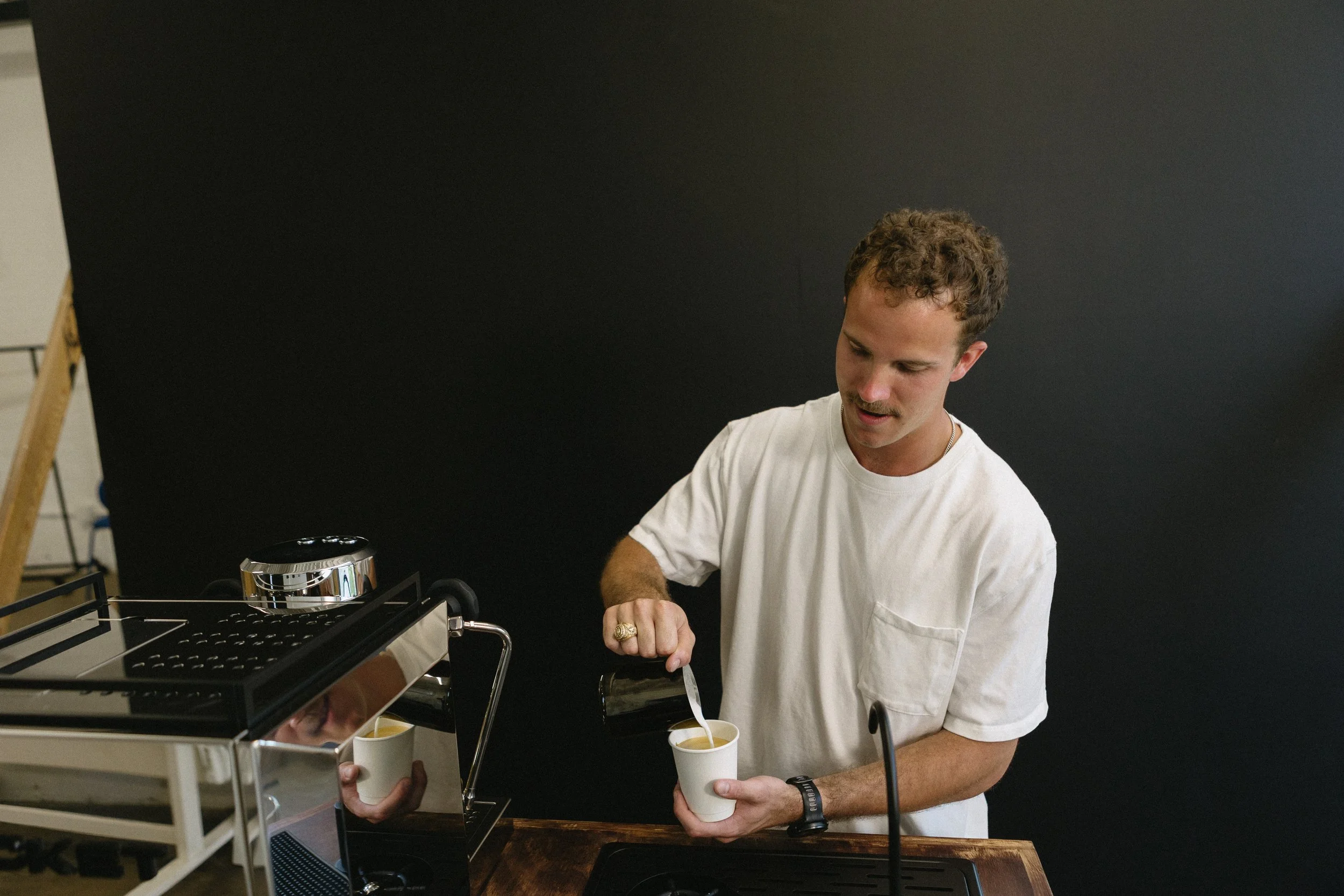 A man wearing a white t-shirt is pouring a beverage from a dispenser into a white cup, with another person holding a cup. They are in front of a black background, possibly at a catering or coffee station.