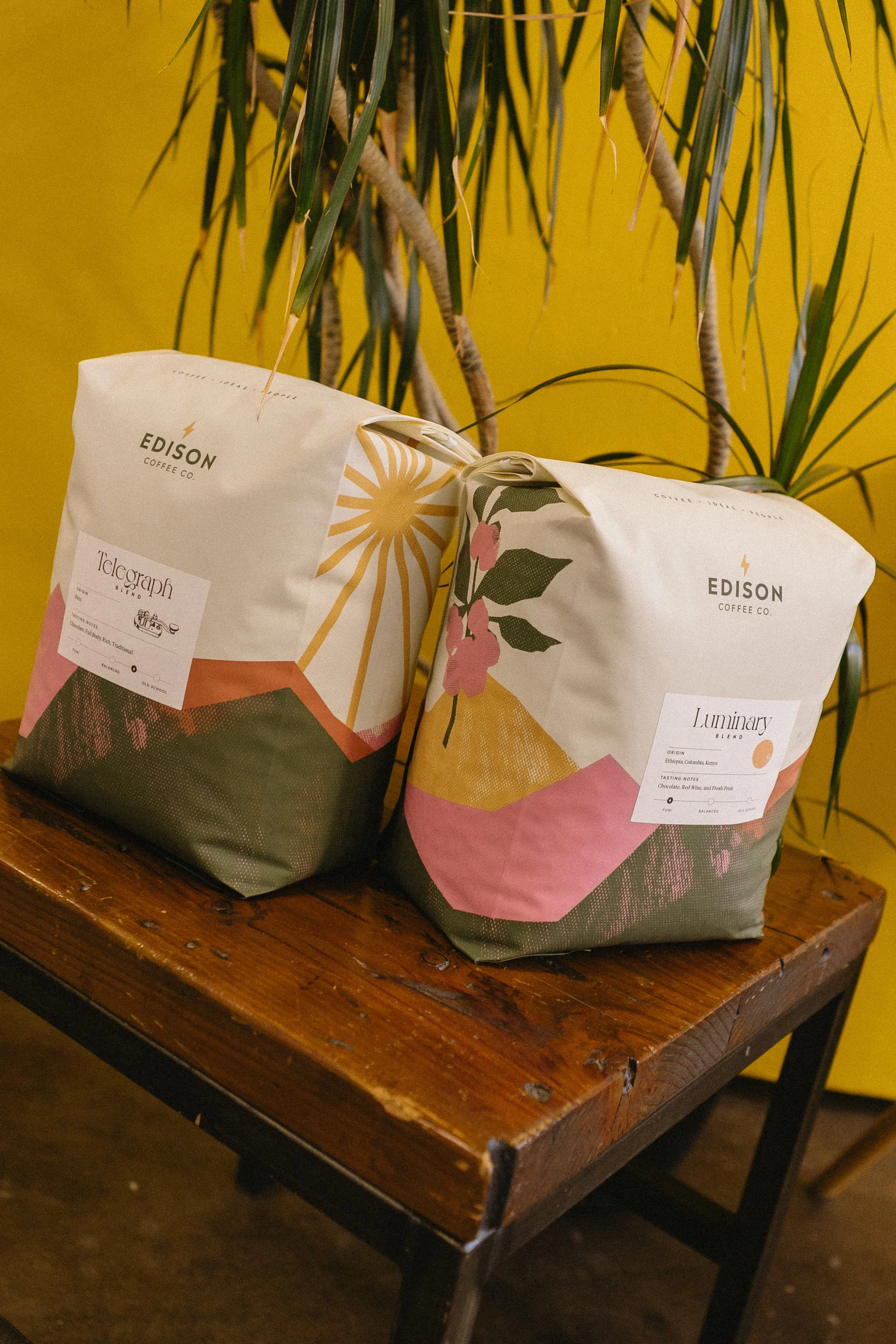 Two bags of coffee from Edison Coffee Co. placed on a wooden table with a yellow wall and a large leafy plant in the background.