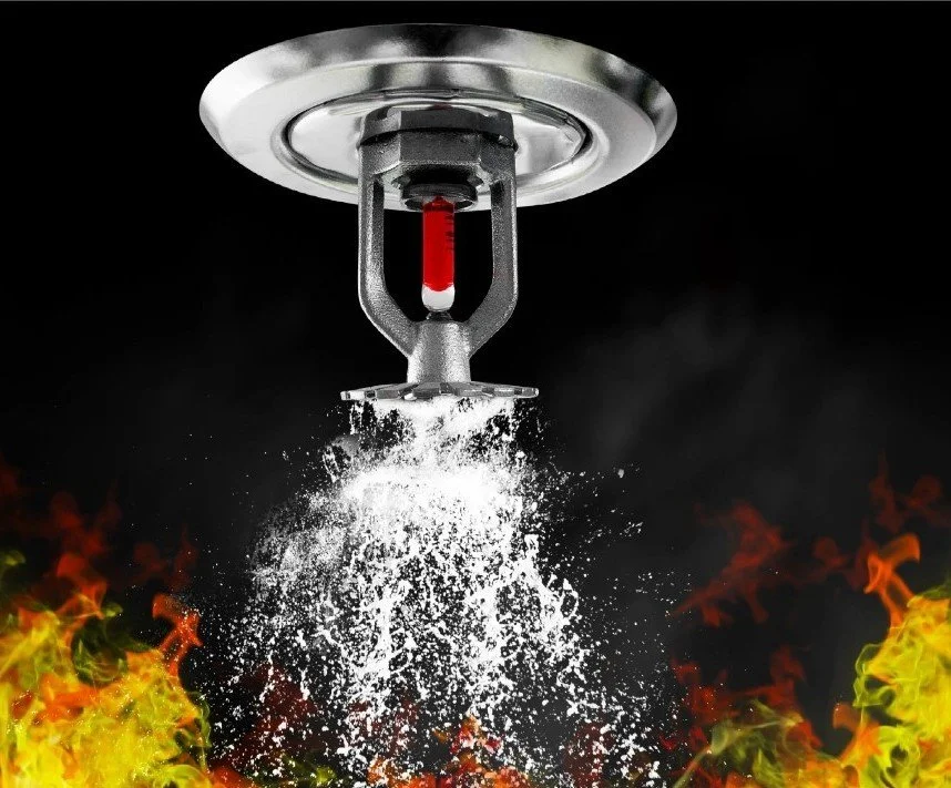 Fire sprinkler head releasing water above flames with a red sprinkler indicator in a black background.