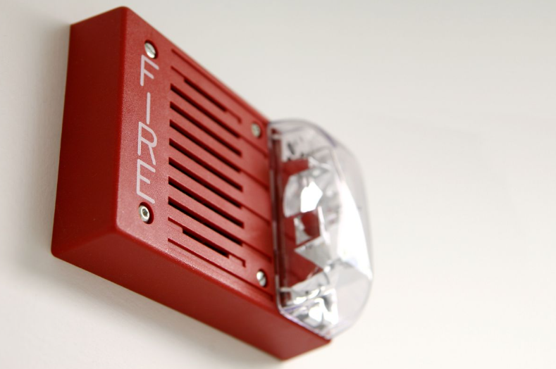 A red fire alarm with a clear plastic cover mounted on a white wall.