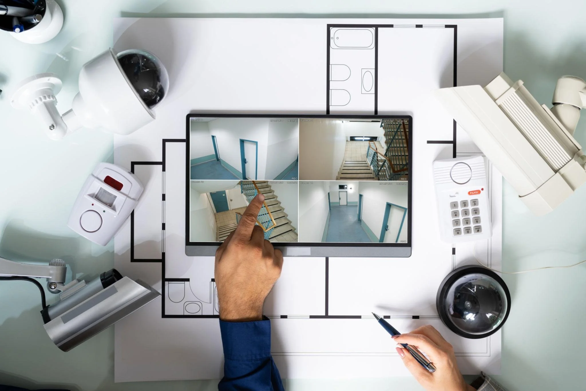 Overhead view of a person pixelating a staircase on an architectural floor plan using a tablet. Surrounding are a security camera, a fire alarm, a pen, a rotary dial phone, and other electronic devices.