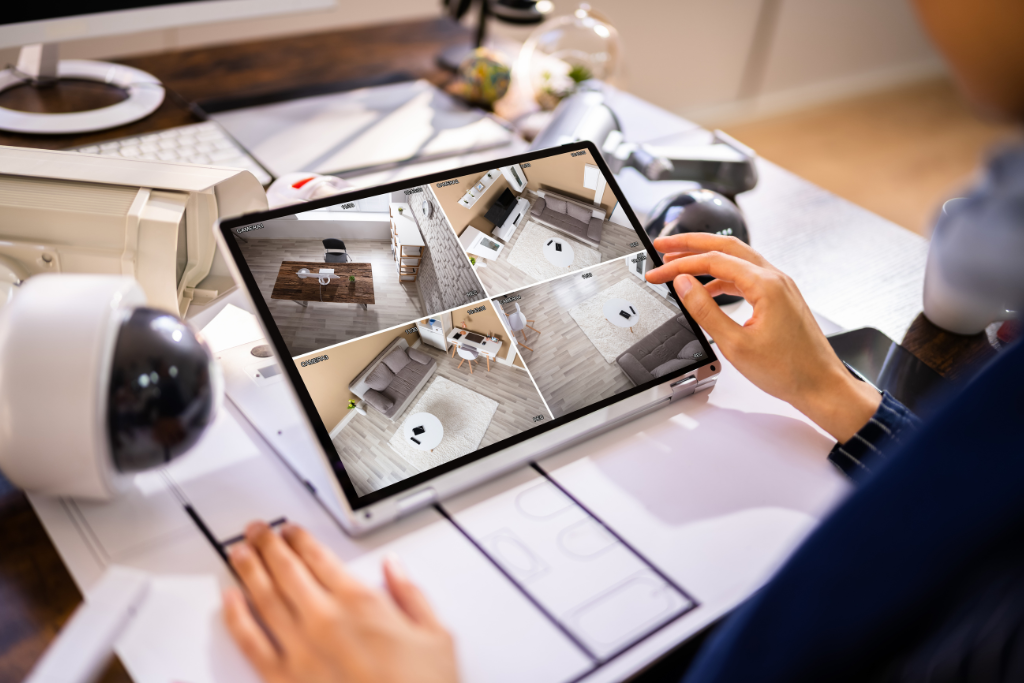 Person monitoring security camera feeds on a tablet in an office or home setting, with various tech devices and papers on the desk.