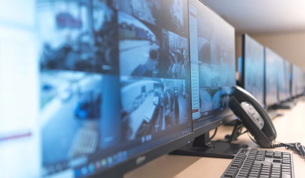 Multiple computer monitors displaying security camera footage, with a telephone and keyboard on the desk.